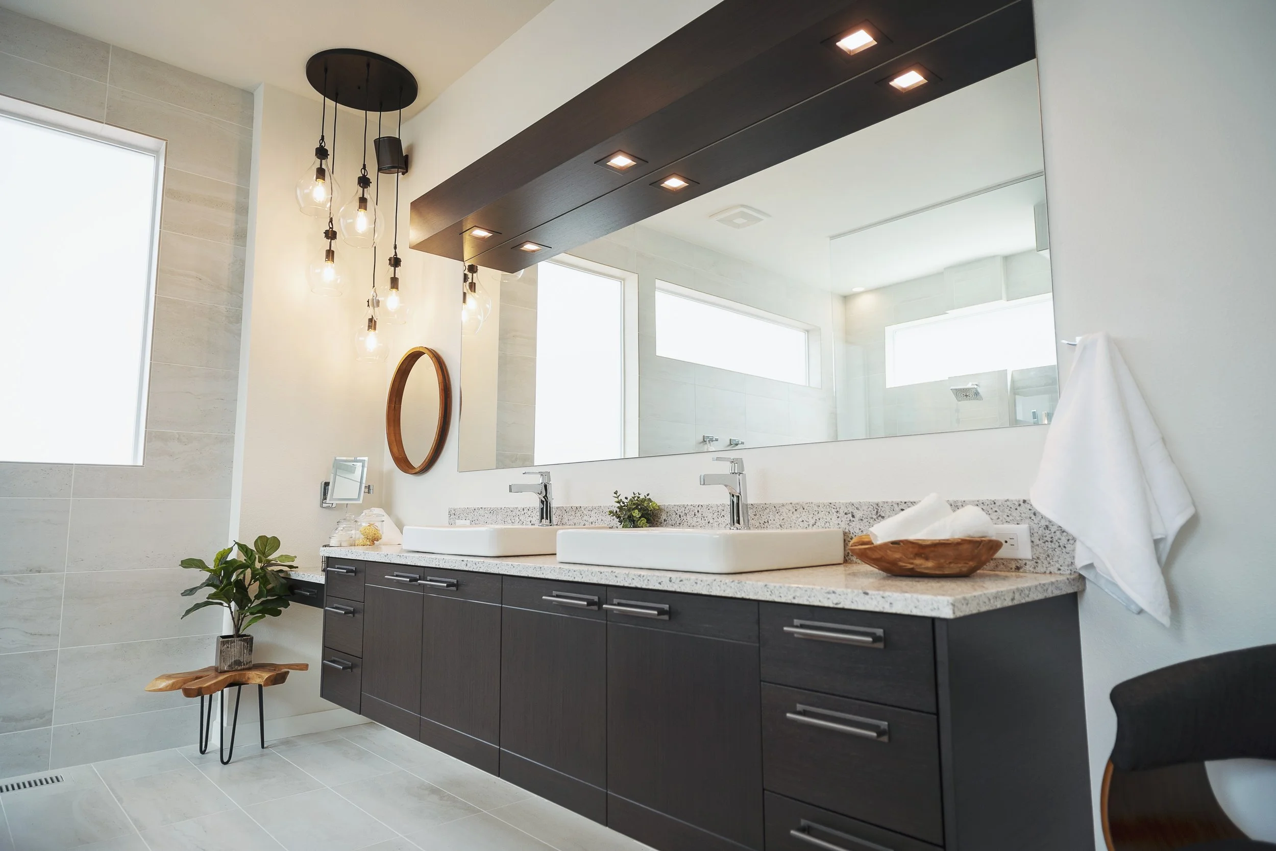 Modern bathroom with a floating dark wood vanity, a large mirror, double sinks, and contemporary lighting fixtures, with a plant on a small wooden table and large windows allowing natural light.
