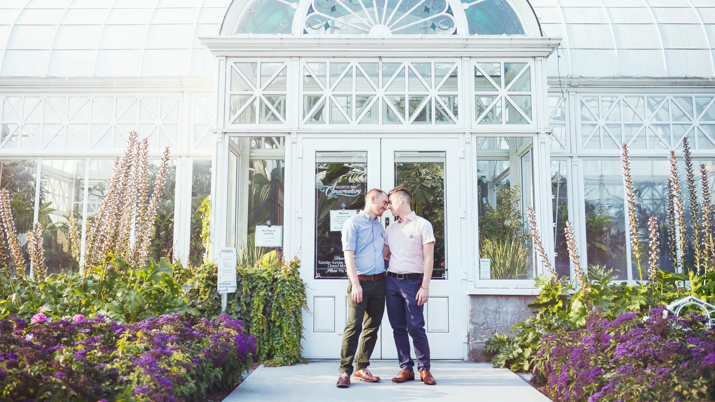 Two men stand close together outside a glass conservatory, touching foreheads in an affectionate pose. The conservatory is surrounded by blooming purple flowers and lush greenery. The glass building has a white framework and a sign on the door that r