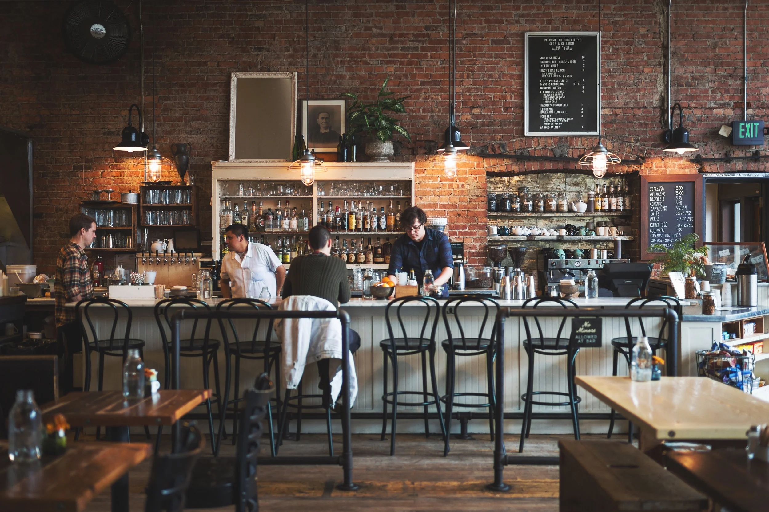 Inside a cozy brick-walled cafe with a barista preparing drinks behind the counter. There are chairs and tables in the foreground, and various bottles and a menu board on the wall.