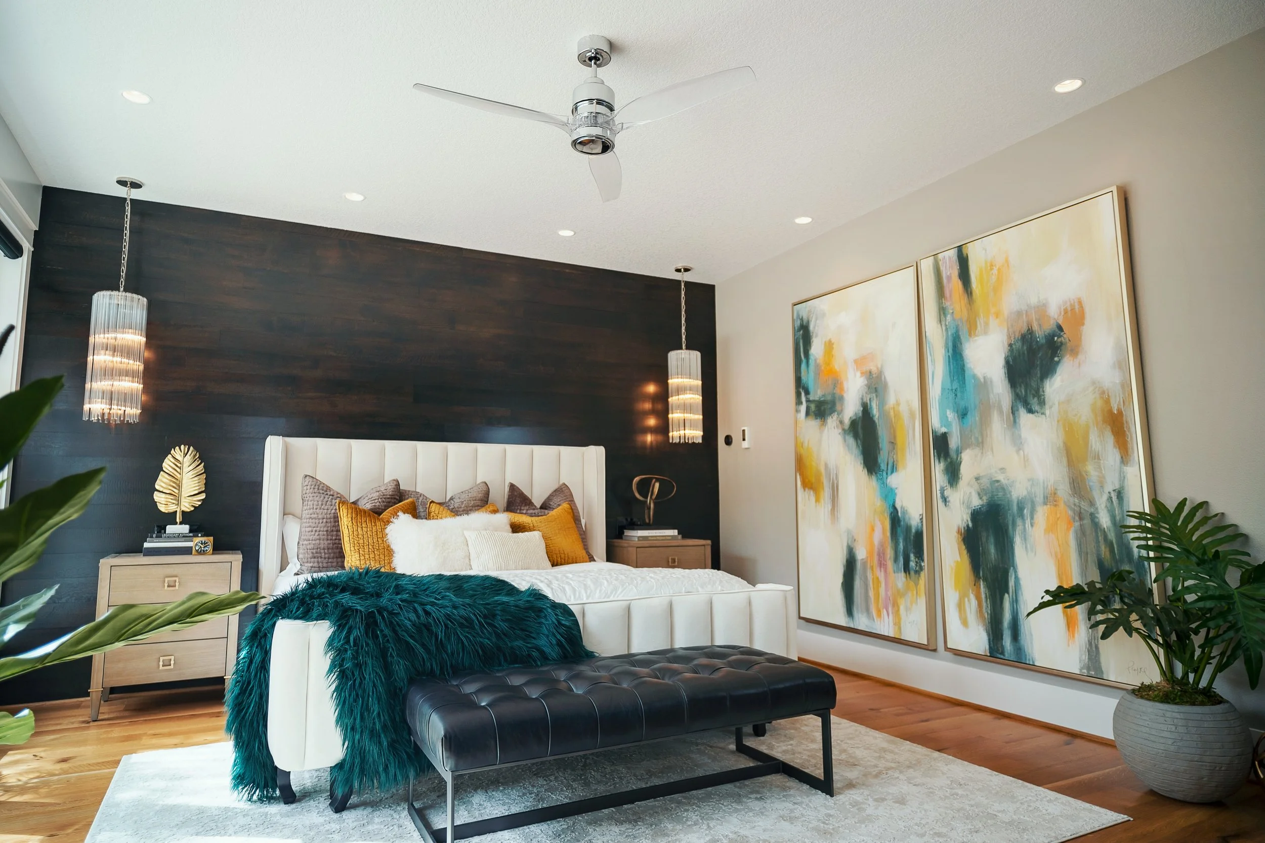 Modern bedroom with white upholstered bed, colorful pillows, and a teal throw. Dark wood accent wall with two hanging pendant lights, large abstract paintings, and lush green plants. Ceiling fan and natural light.