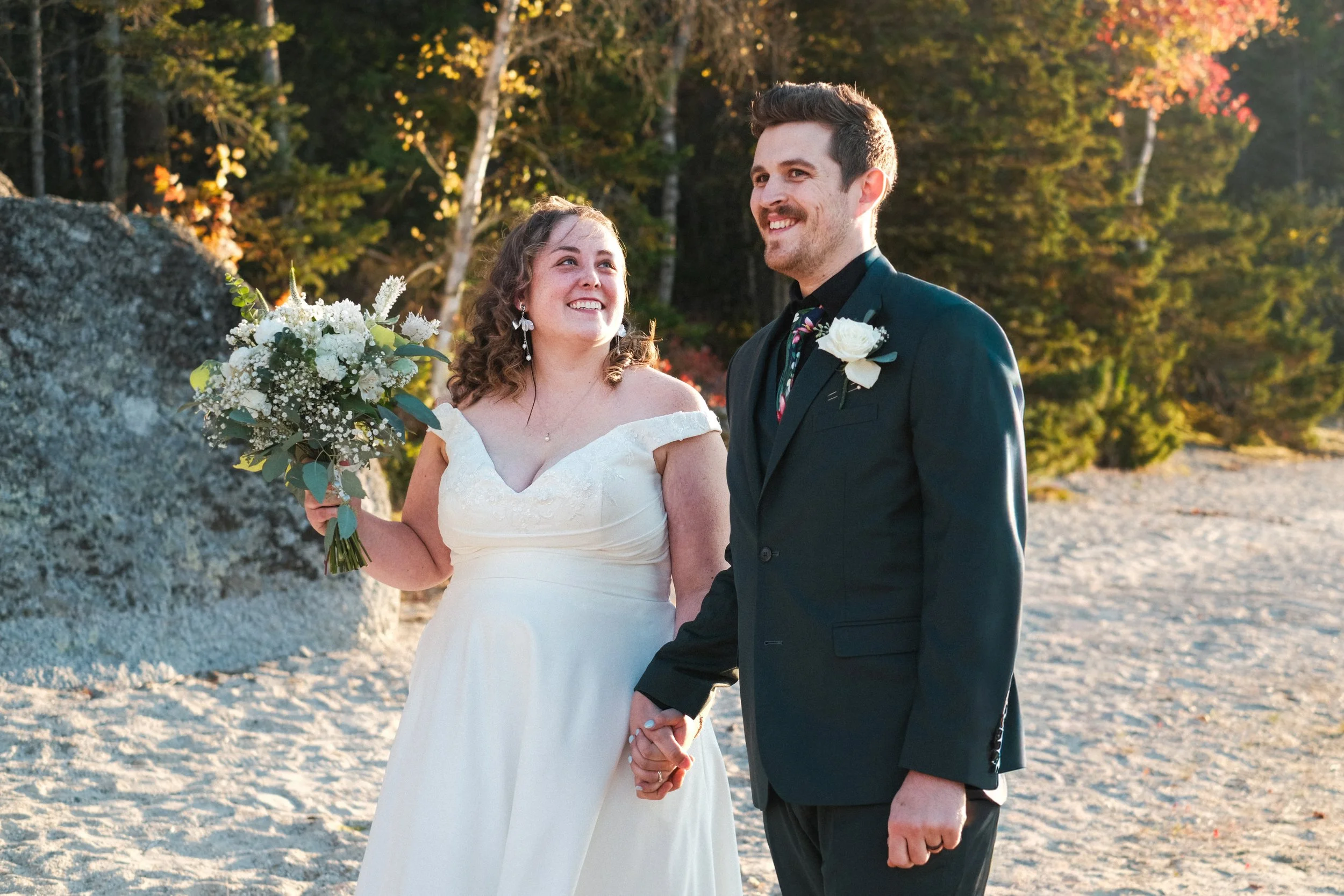 A bride and groom holding hands outdoors during their wedding, smiling and looking at each other, with autumn trees in the background.