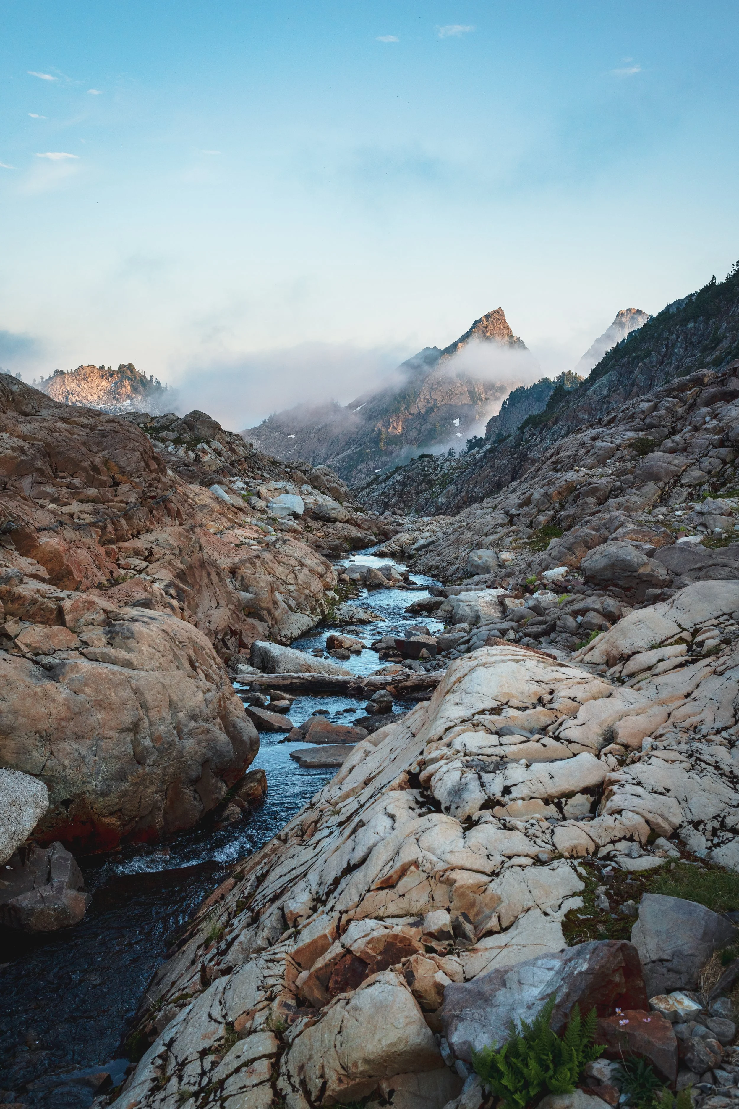 A mountain stream flows through rocky terrain with large boulders and patches of greenery, with snow-capped peaks and mist in the background under a partly cloudy sky.