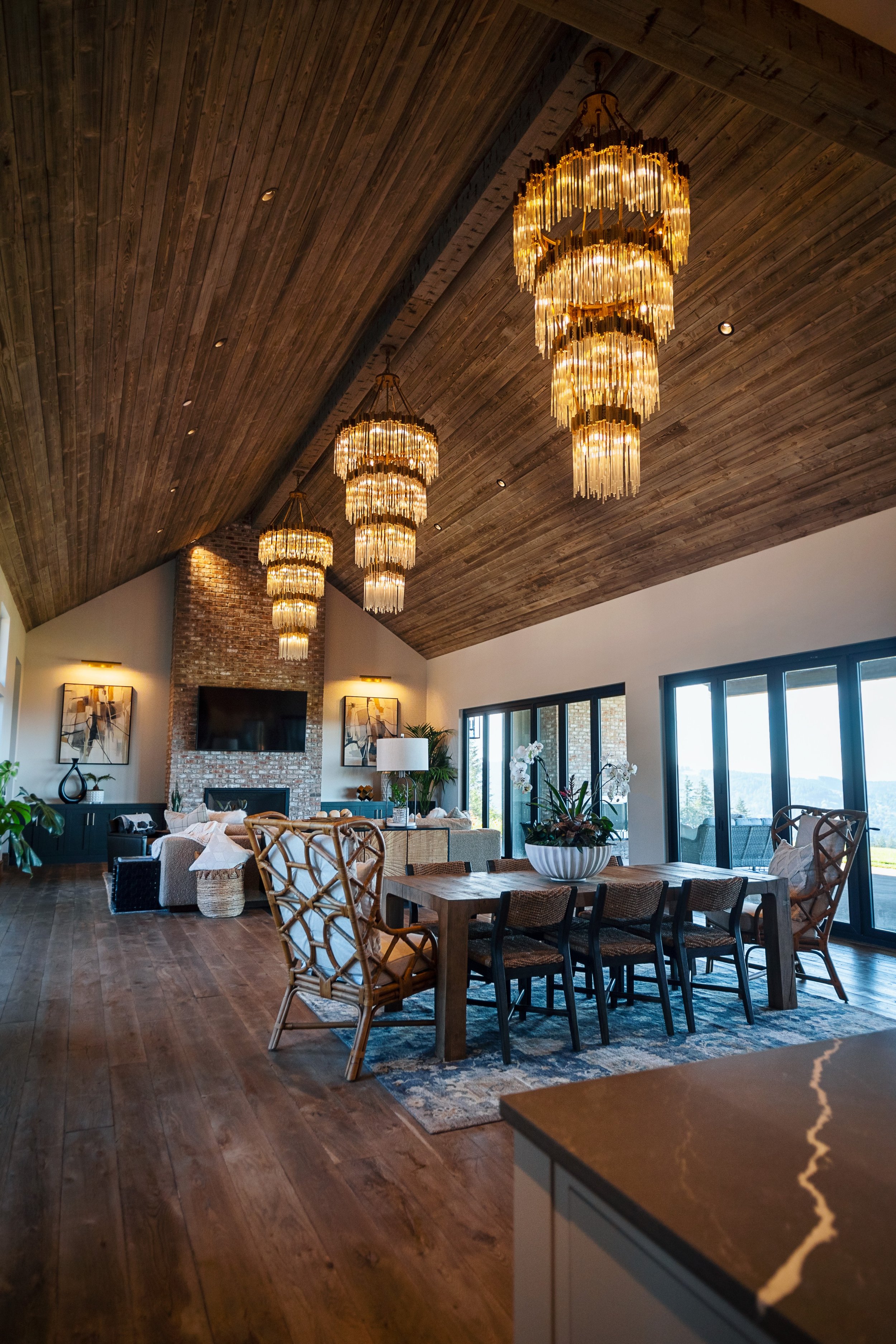 Interior view of a living and dining area with a high wooden vaulted ceiling, large hanging chandeliers, a brick fireplace with a flat-screen TV, artwork on the walls, a large dining table with chairs, and a seating area with sofas and plants near sl