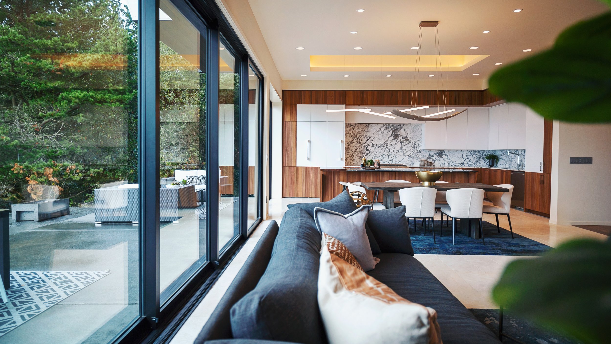 Modern living room with large sliding glass doors leading to outdoor patio, contemporary kitchen with wood and marble accents, dining area with white chairs and a dark wood table, gray sofa with patterned pillows, and green plants.