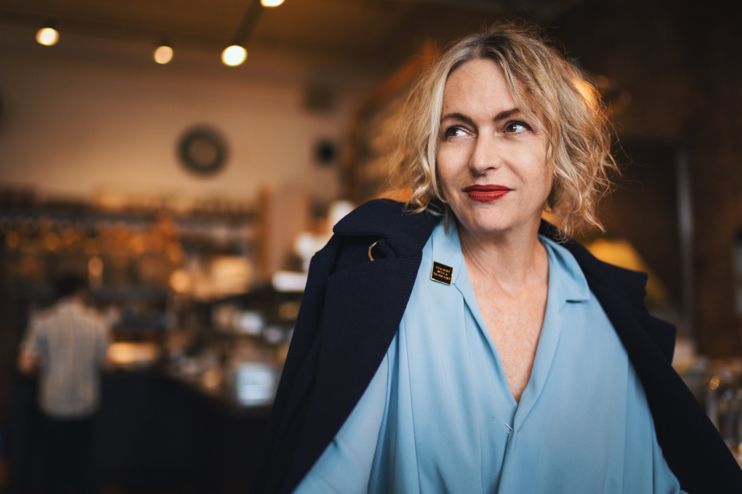 A woman with curly blonde hair and red lipstick, wearing a blue shirt and a dark jacket, inside a warmly lit restaurant or café.