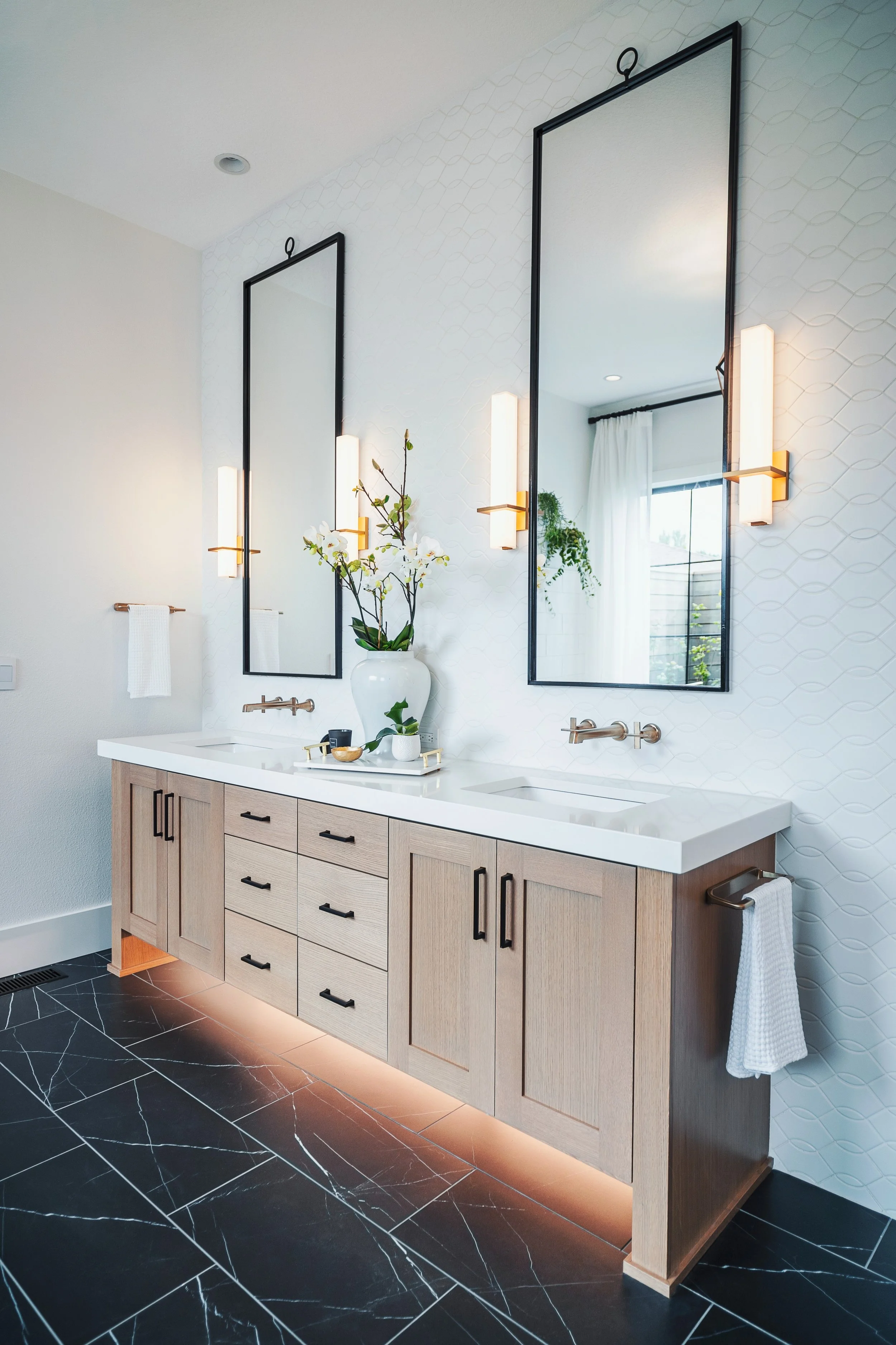 Hotel or bathroom vanity with a double mirror, modern lighting fixtures, a wood cabinet with multiple drawers, decorative items including a vase with flowers, and black marble floor tiles with white veining.