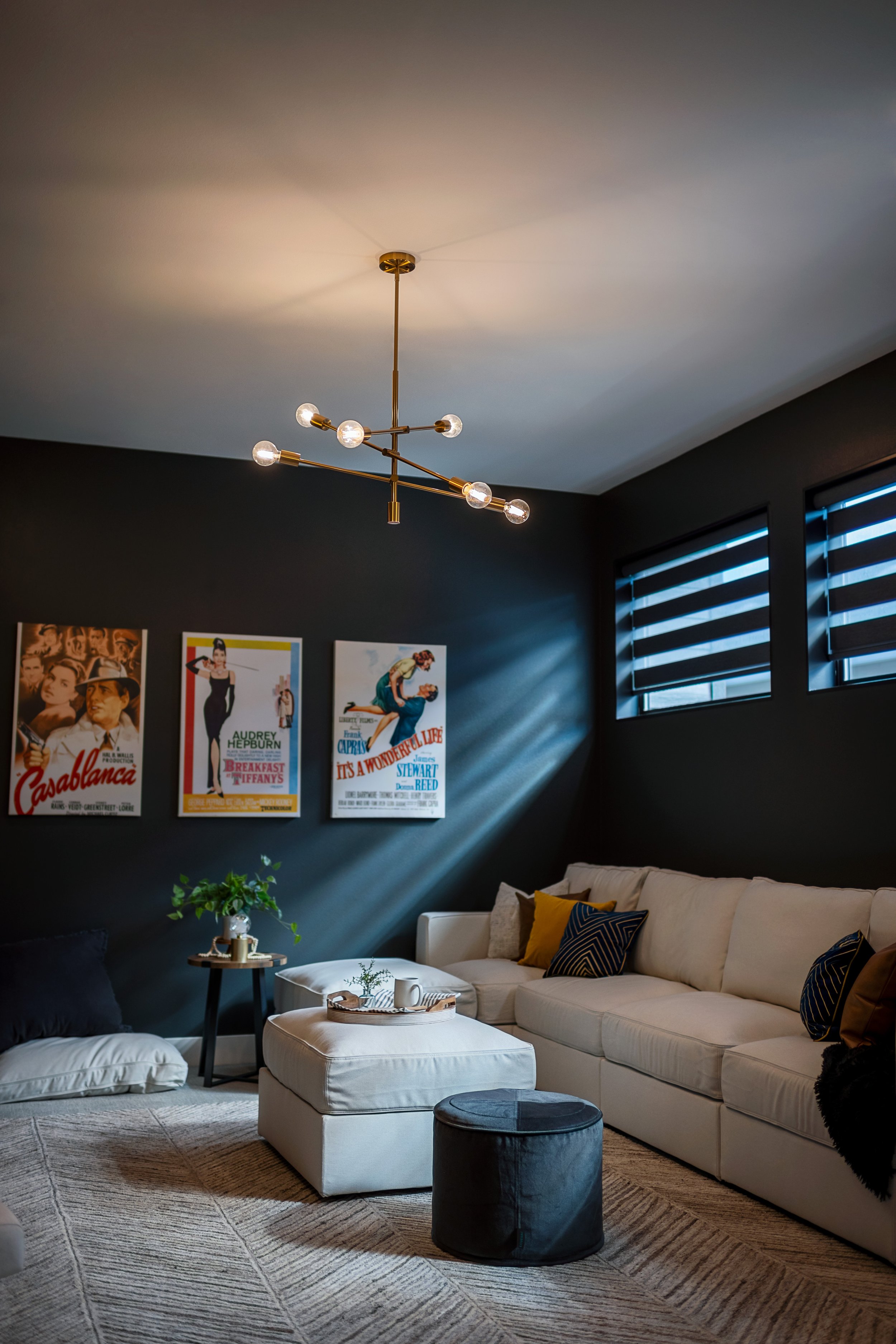 Living room with a modern chandelier, a beige sectional sofa with black, yellow, and patterned pillows, and wall art posters. There is a small side table with a plant and a white ottoman in front of the sofa. The walls are dark, and windows have stri