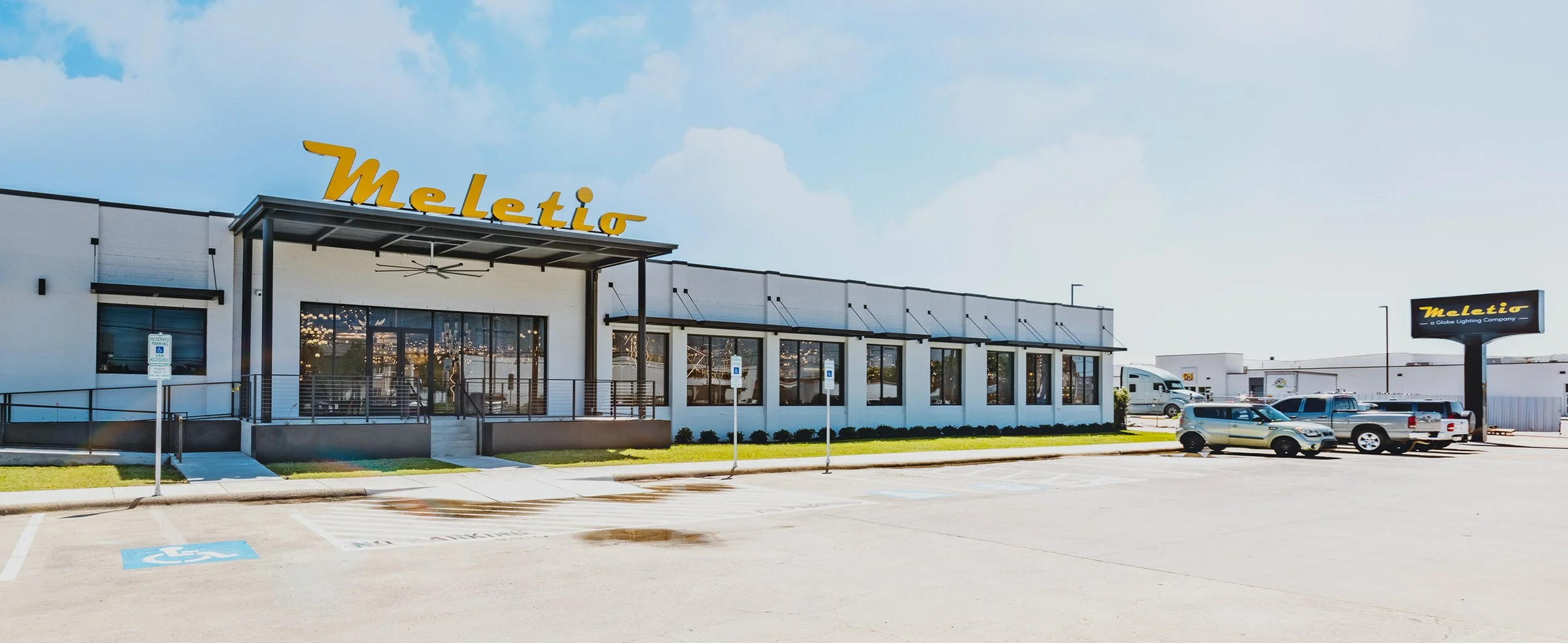 Exterior of a modern commercial building with a sign that reads 'Meletio' and parking lot in front with several parked cars
