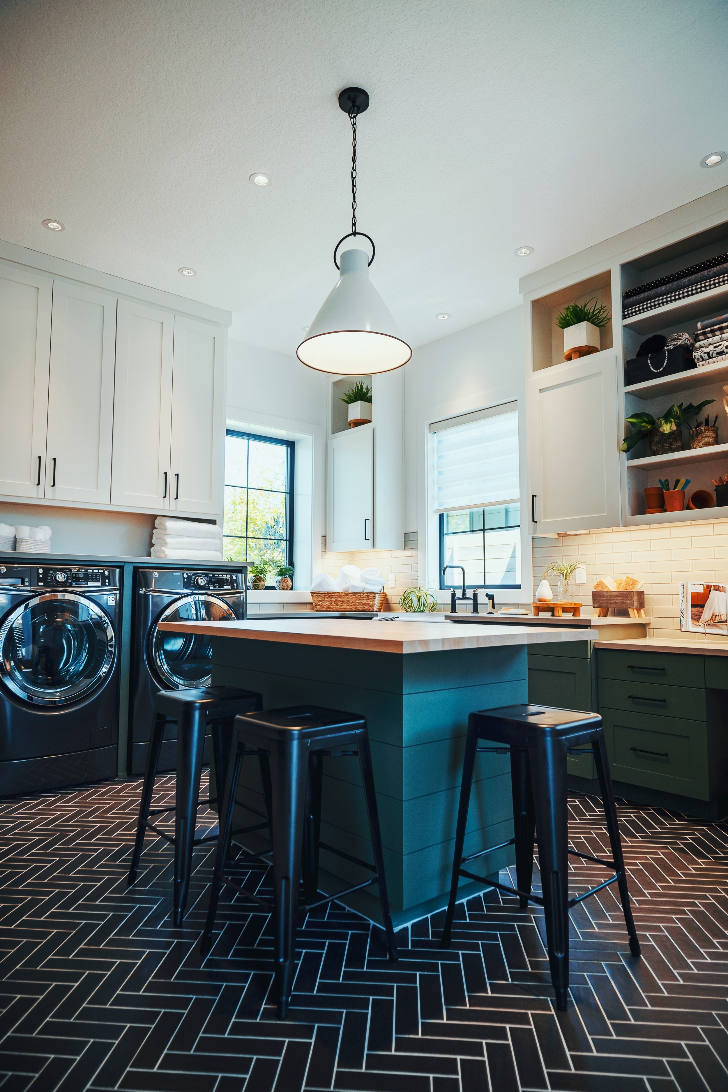 Modern kitchen with white cabinets, a kitchen island, black bar stools, and black and white patterned tile flooring. There are large windows, green plants, and open shelves with kitchen items.