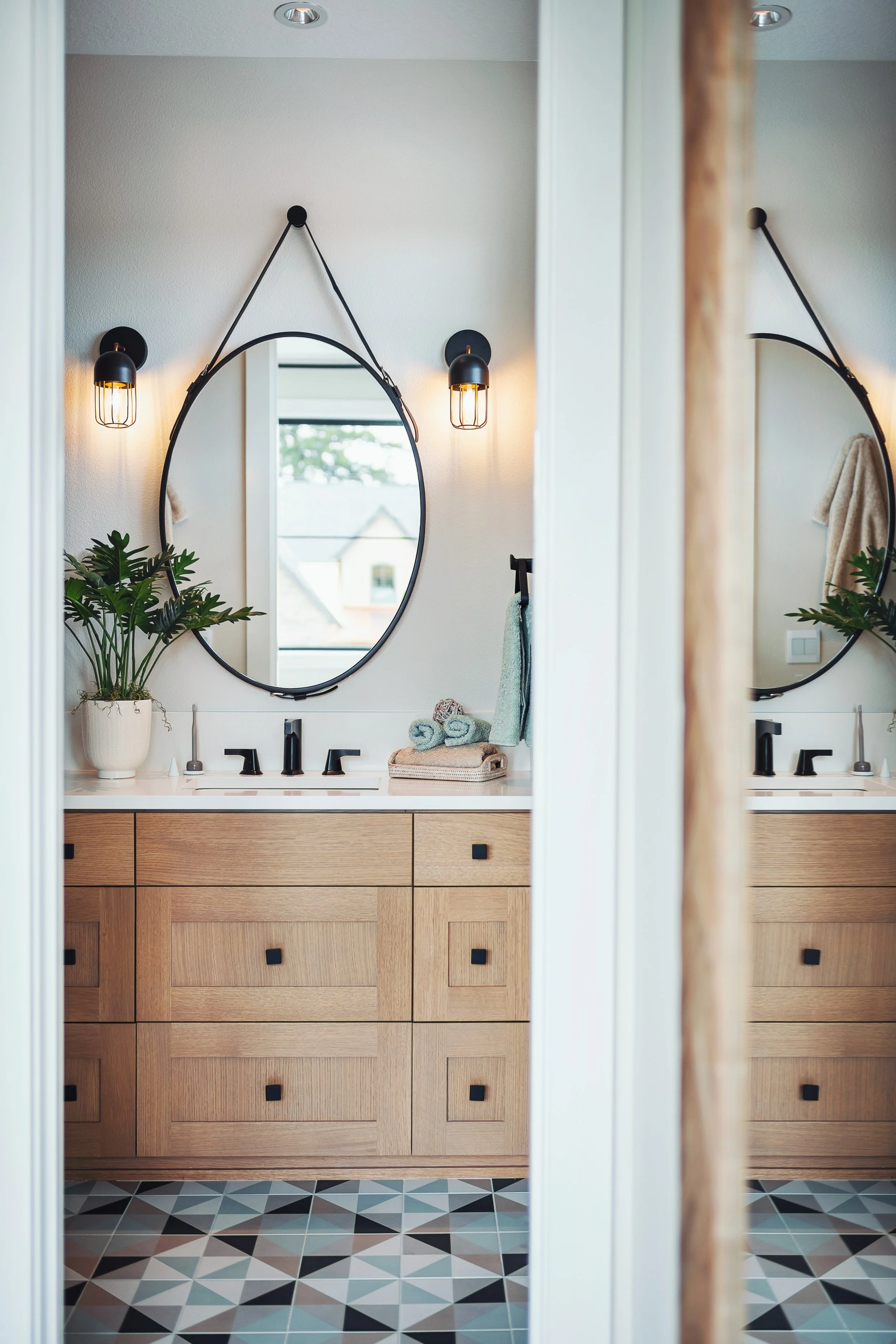 A modern bathroom with a wooden vanity, black faucets, an oval mirror, and black wall sconces with exposed bulbs. There is a potted plant, towels, and a window reflected in the mirror, with geometric patterned tiles on the floor.