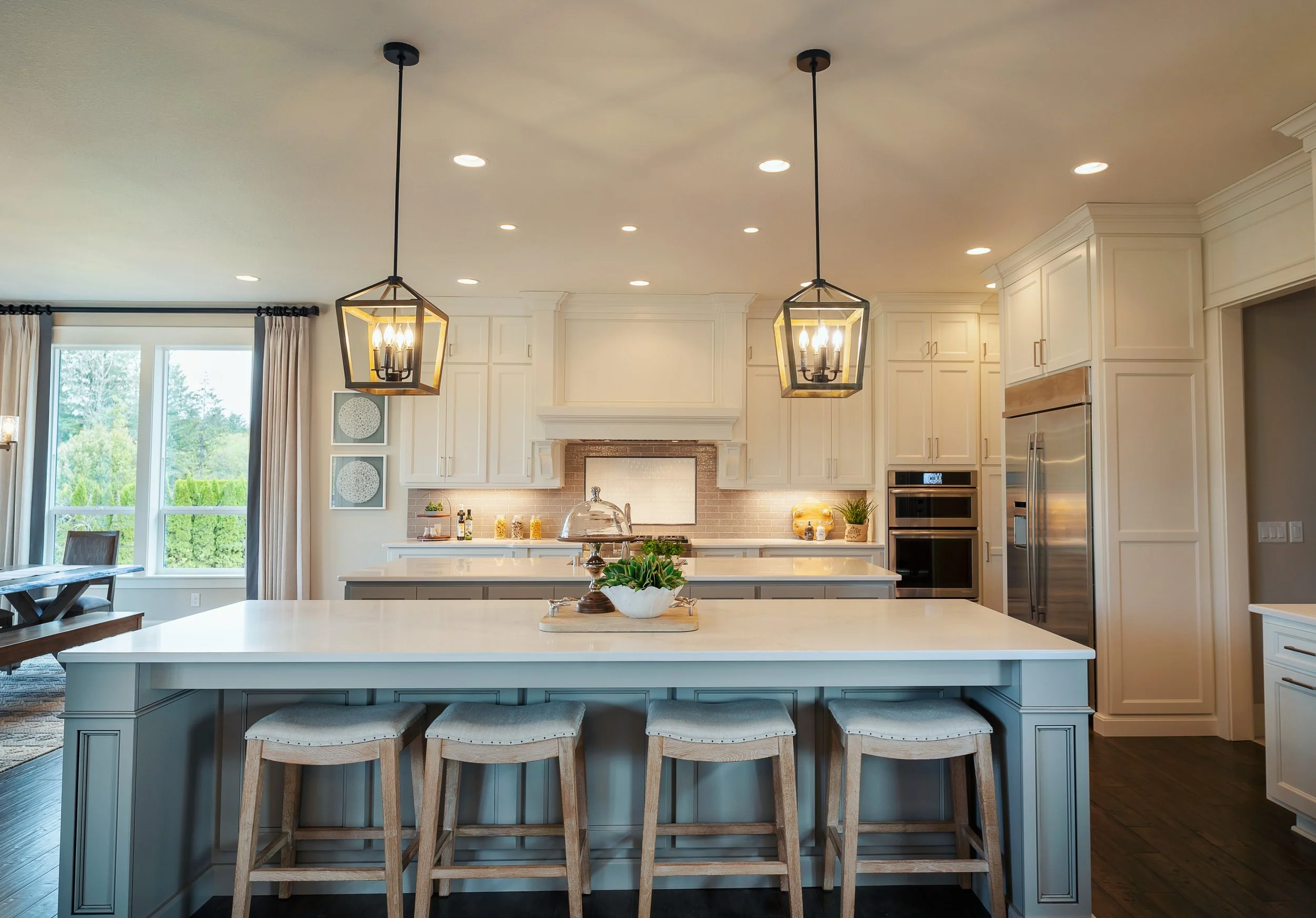 Modern kitchen with white cabinets, a large kitchen island with gray base and white countertop, three wooden barstools with upholstered seats, two pendant lights hanging from the ceiling, a window with curtains, and a view of greenery outside.