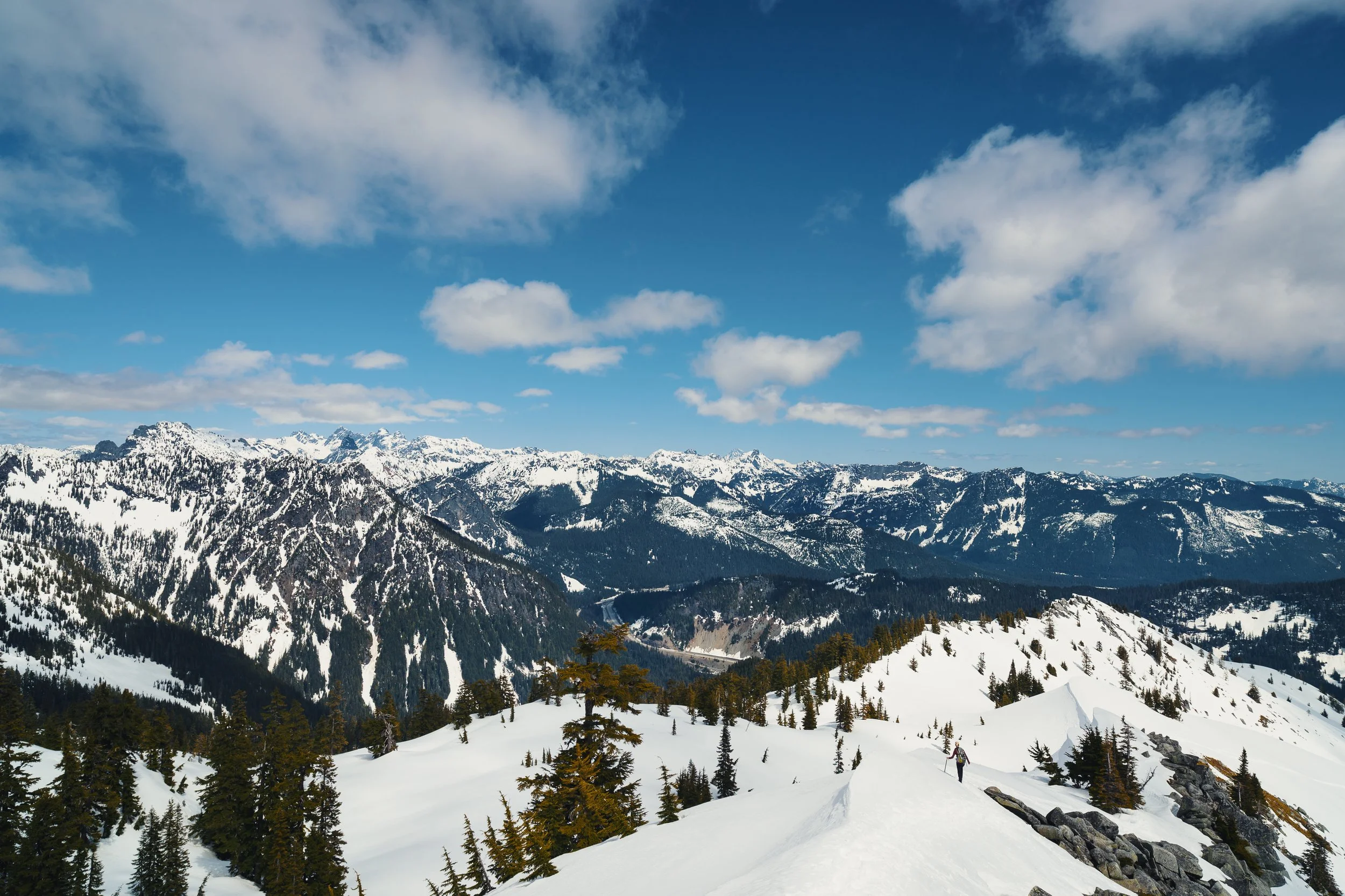 Snow-covered mountains under a partly cloudy blue sky with a few hikers or skiers in the distance on a snow trail.