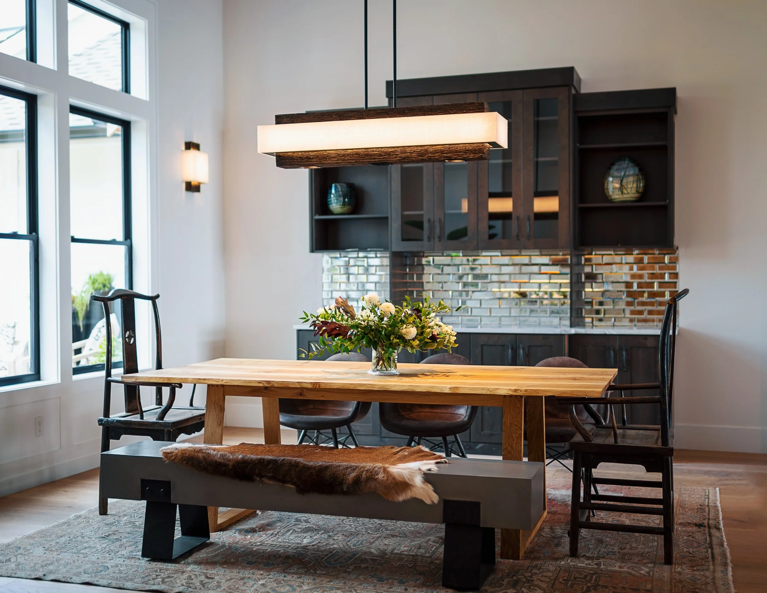 Interior of a modern dining room with large windows, a wooden dining table with a floral centerpiece, black chairs, a patterned rug, and a dark wooden cabinet with decorative vases, illuminated by a rectangular pendant light and wall sconces.