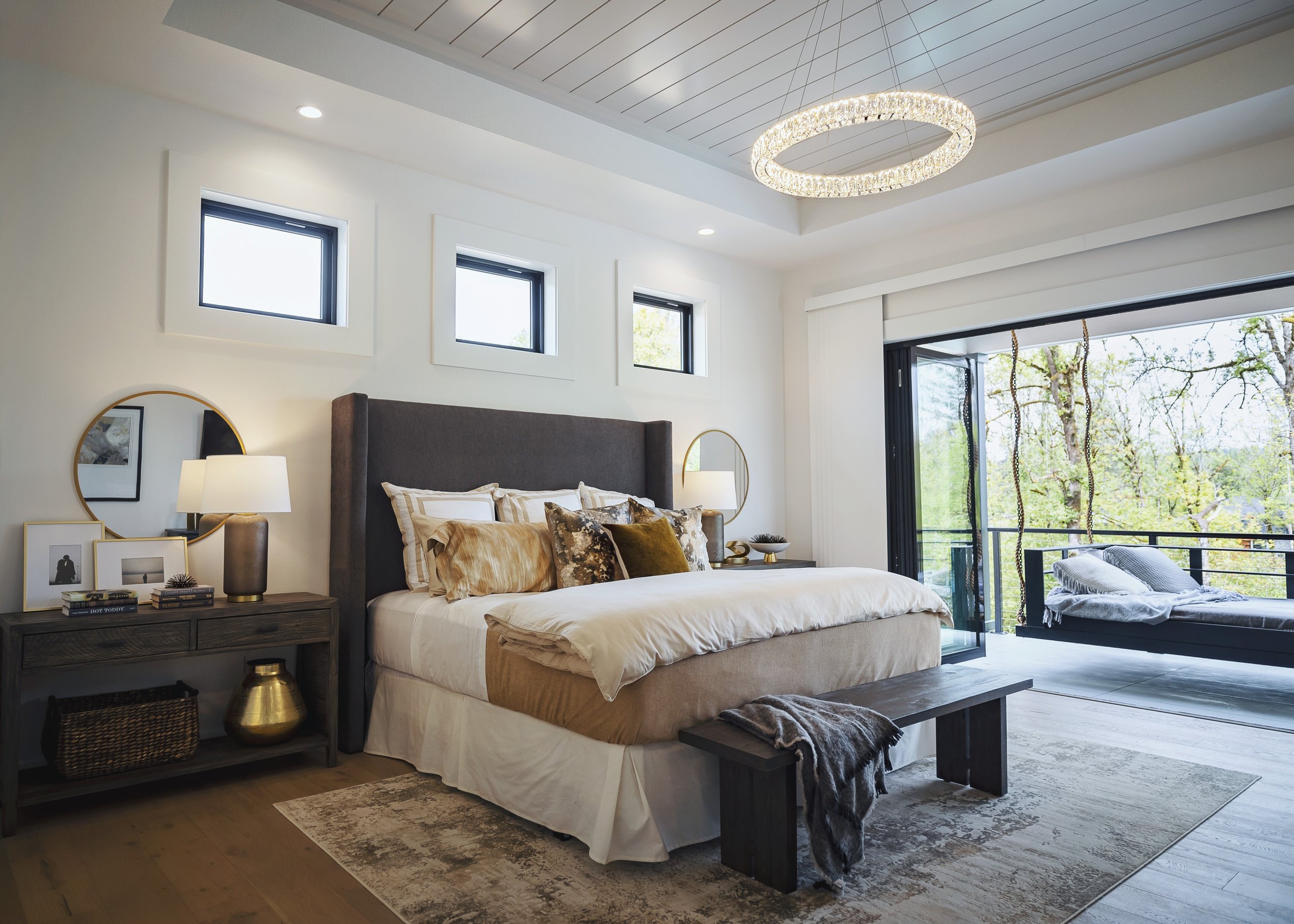 A modern bedroom with a large bed, dark upholstered headboard, and a nightstand with a round mirror, lamps, and decorative items. There are three small windows on the wall and a sliding glass door leading to a balcony with outdoor furniture and trees
