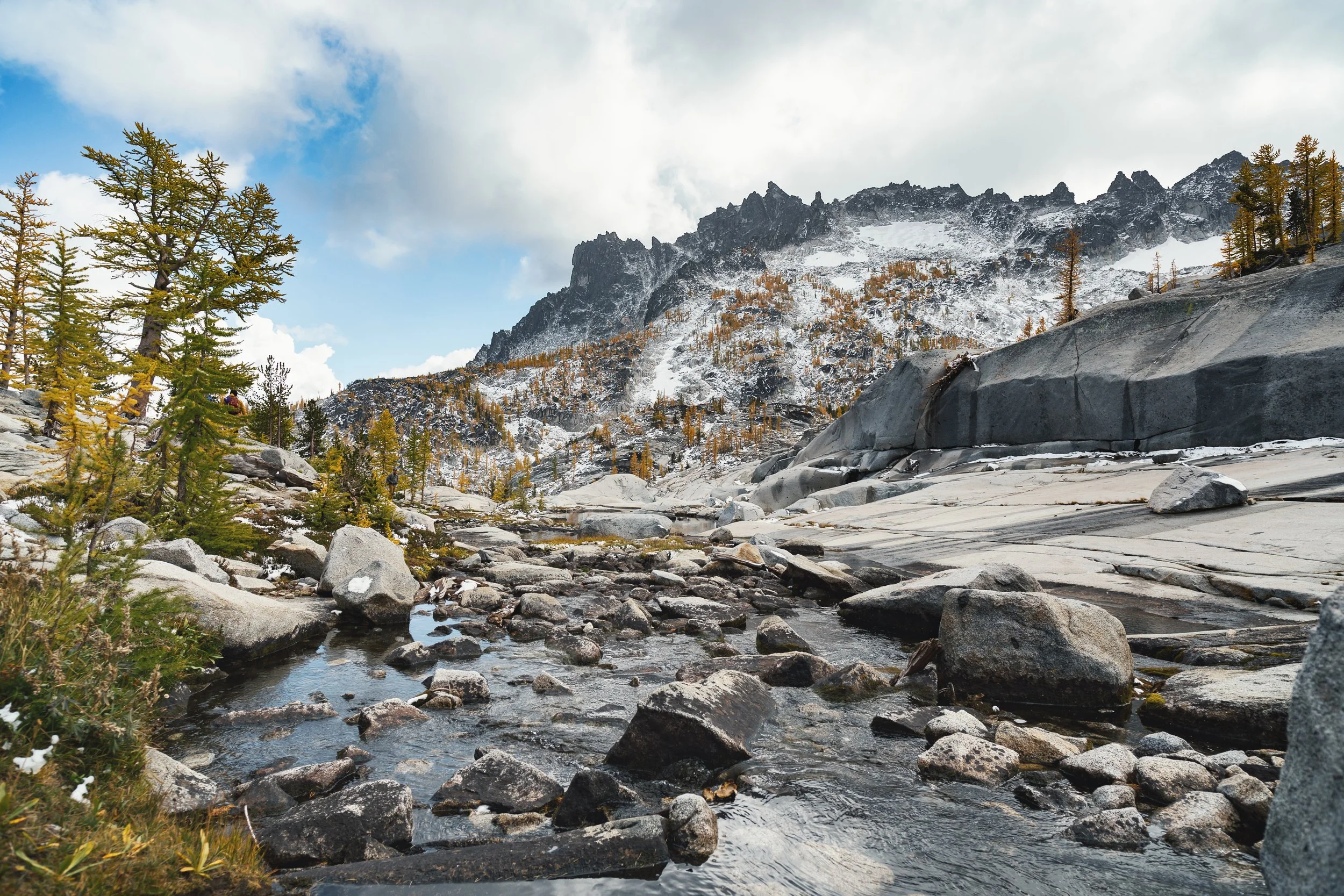 A mountainous landscape with a snow-capped mountain range in the background, scattered trees on the hillside, and a rocky stream in the foreground.