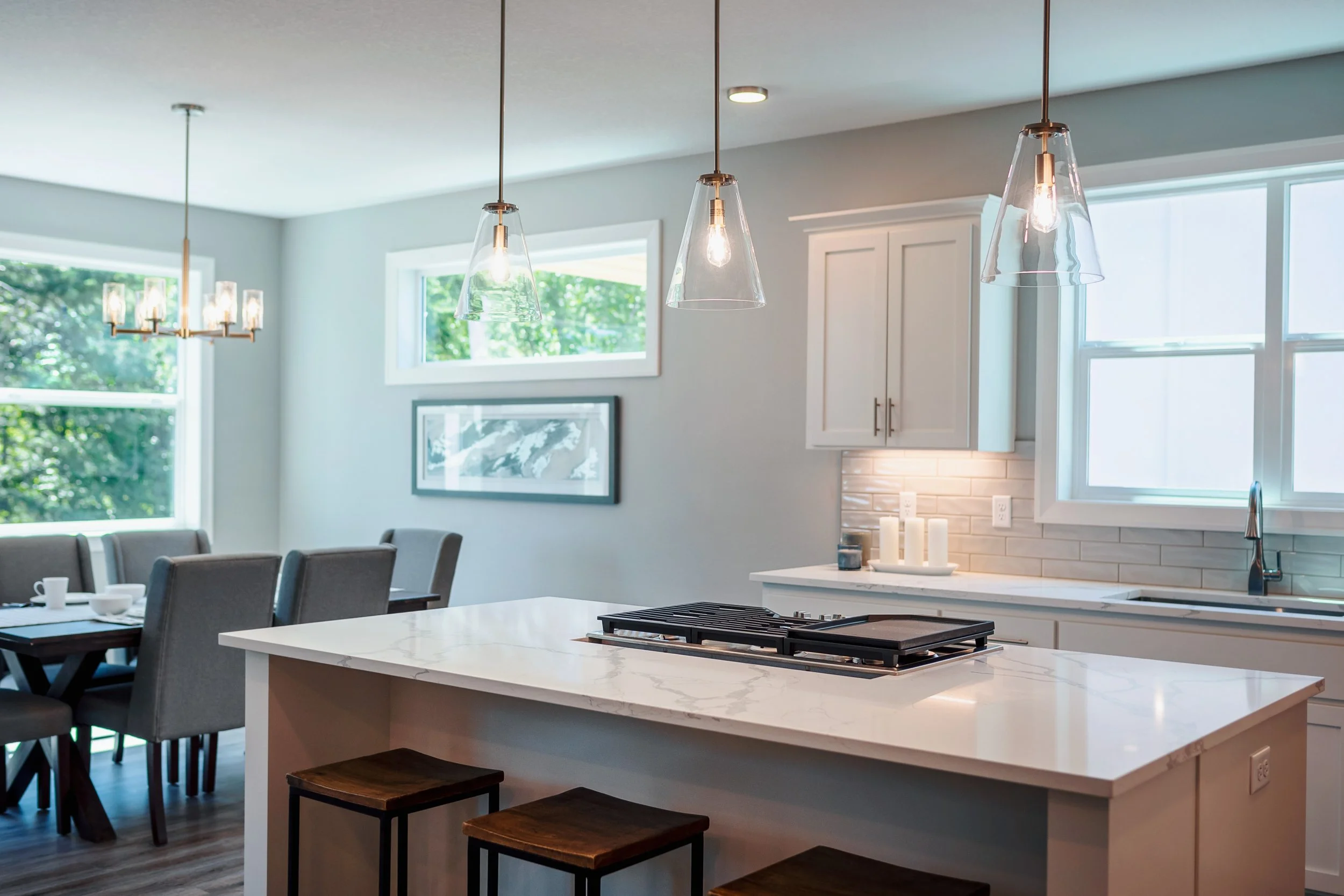 Modern kitchen with white cabinets, a large white island with stools, pendant lights, and two windows showing greenery outside.