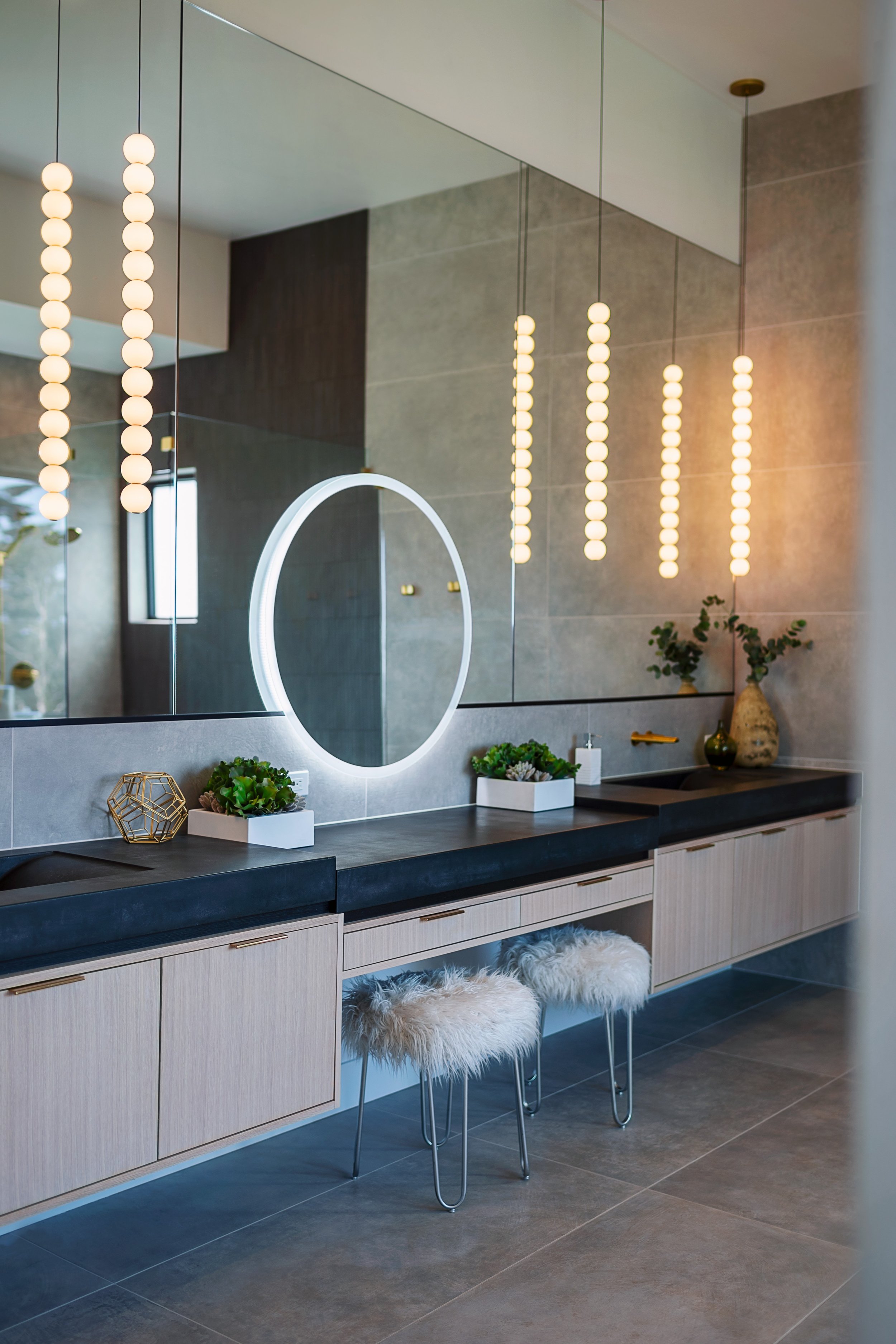 Modern bathroom with a large round backlit mirror, a vanity with two small chairs with fluffy white cushions, plants, and decorative items, illuminated by vertical row of pendant lights and a circular mirror light.