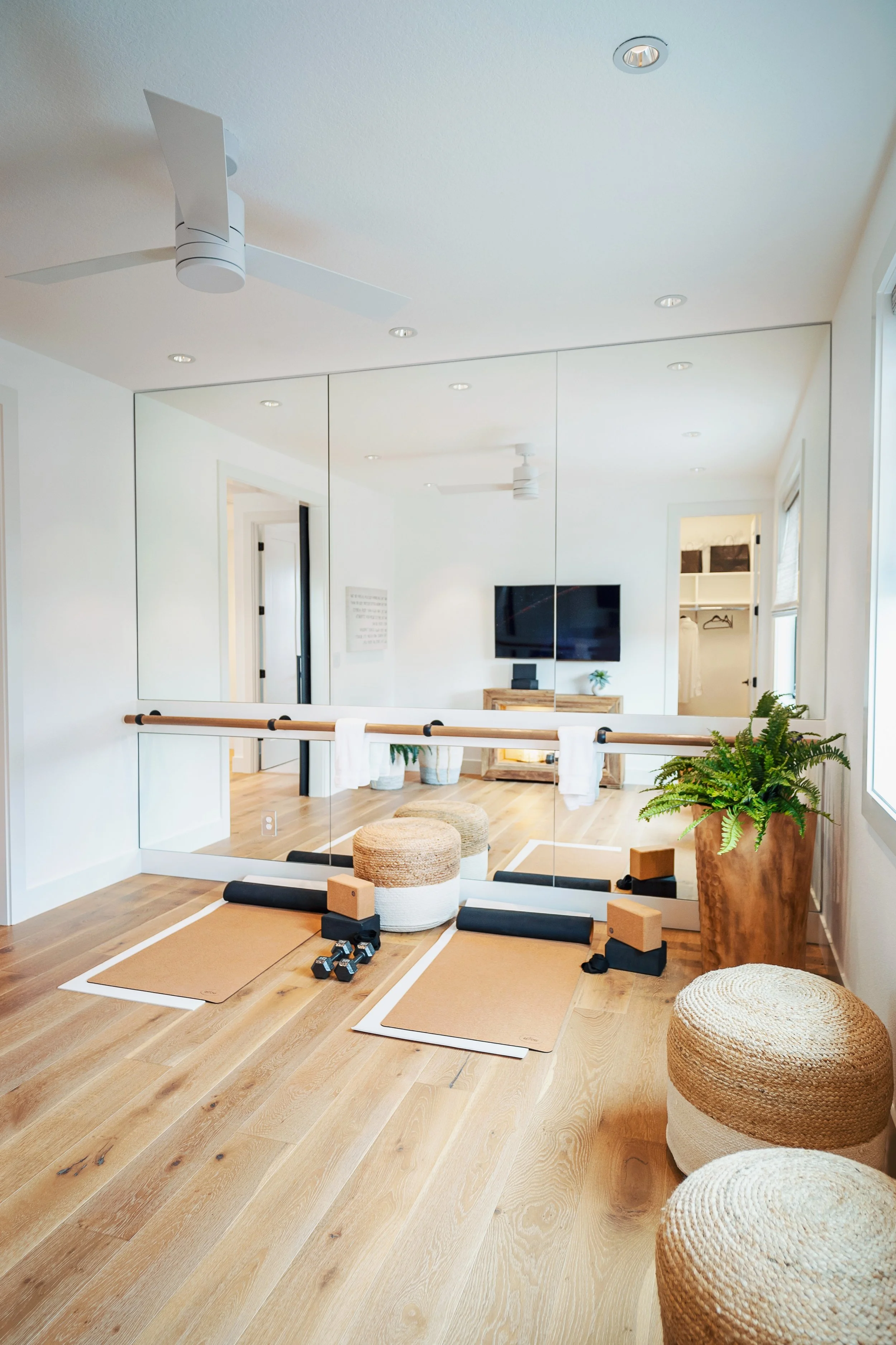 Home yoga or fitness room with large mirror, corner potted plant, exercise mats, dumbbells, foam blocks, bolsters, and woven poufs on light wooden floor. Flat-screen TV on wall, ceiling fan, and recessed lighting.