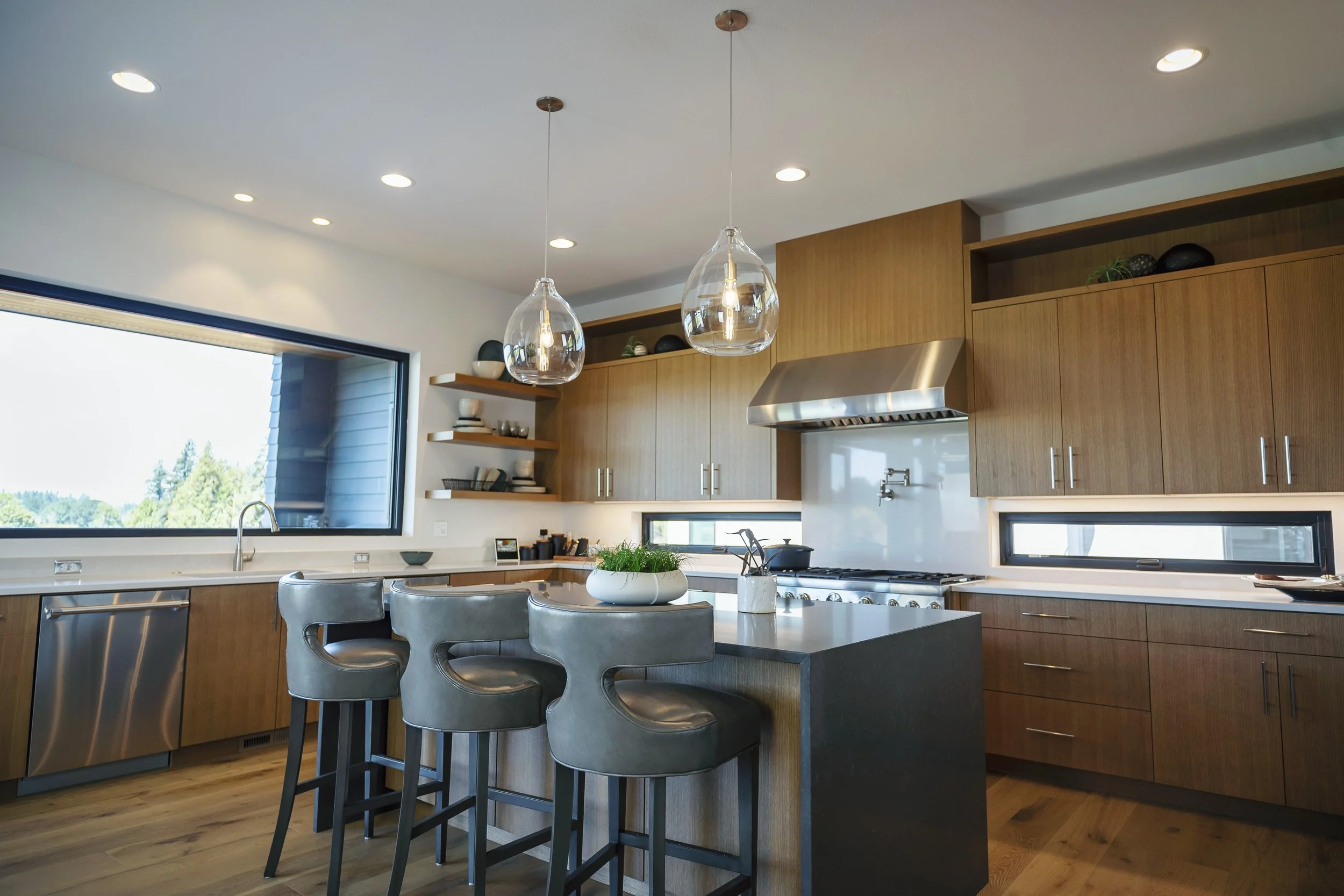 Modern kitchen with wooden cabinets, stainless steel appliances, pendant lights, and large windows showing outdoor trees.