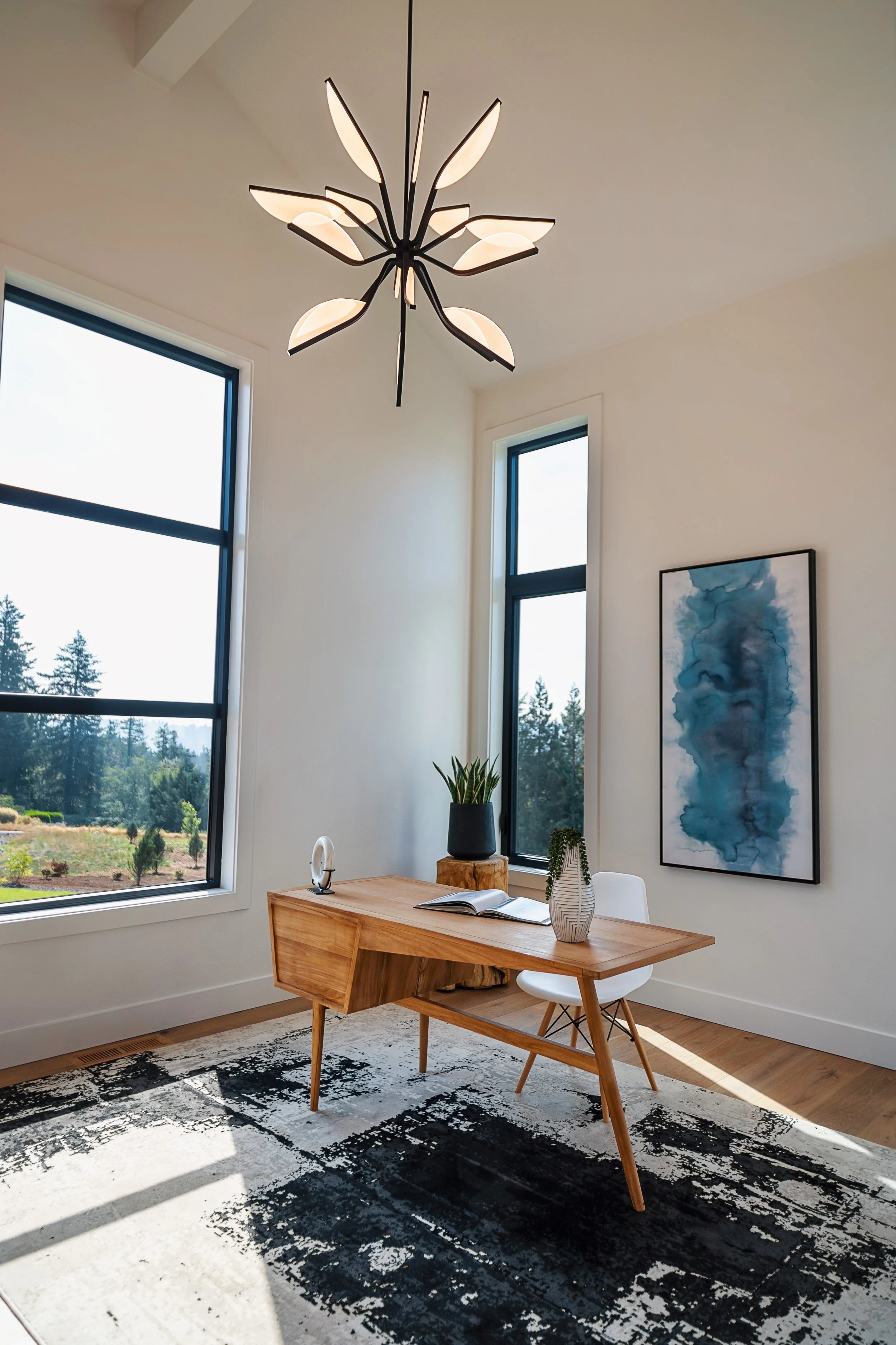 Modern home office with a wooden desk, white chair, potted plants, large windows with outdoor trees, and a blue abstract wall art