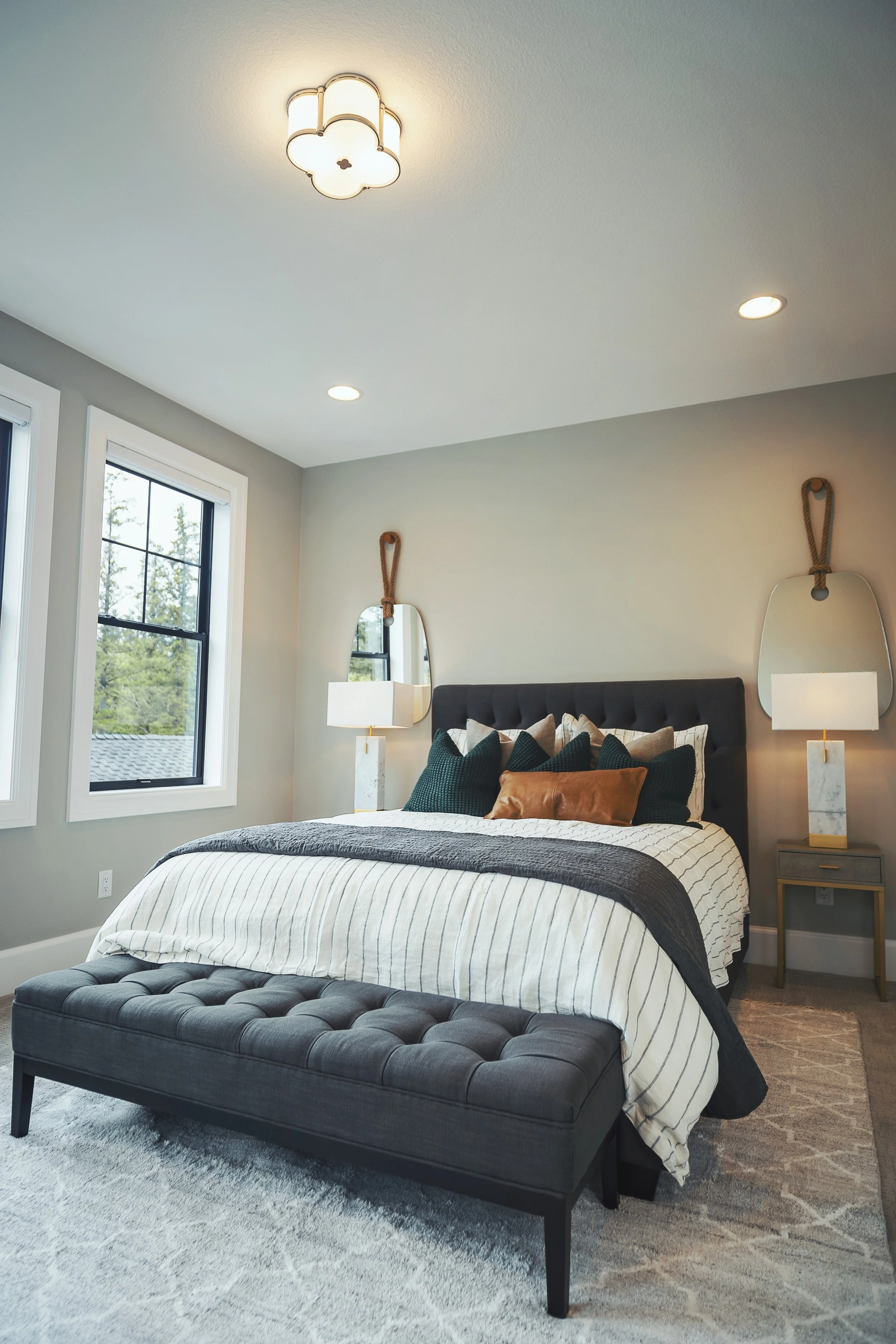 A modern bedroom with a bed that has a black upholstered headboard, white and black striped duvet, and multiple dark green pillows. Two wall-mounted mirrors are above nightstands on either side of the bed, each with a white table lamp. The room featu