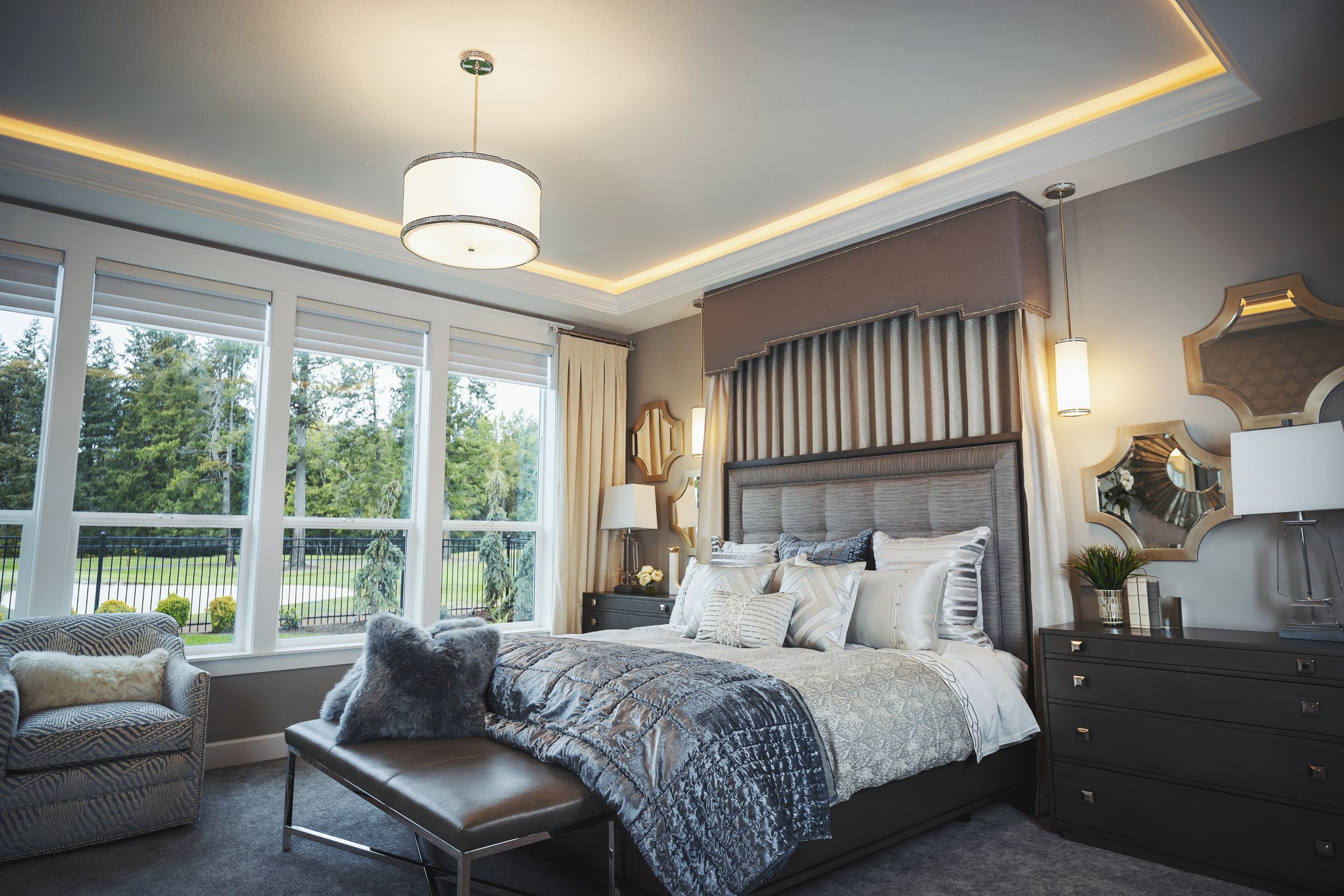 A bedroom with a large bed, decorative pillows, and a gray headboard. There are windows with white blinds and outdoor greenery visible. The ceiling has recessed lighting and a central chandelier. Side tables with lamps, a mirror, and a cozy chair wit