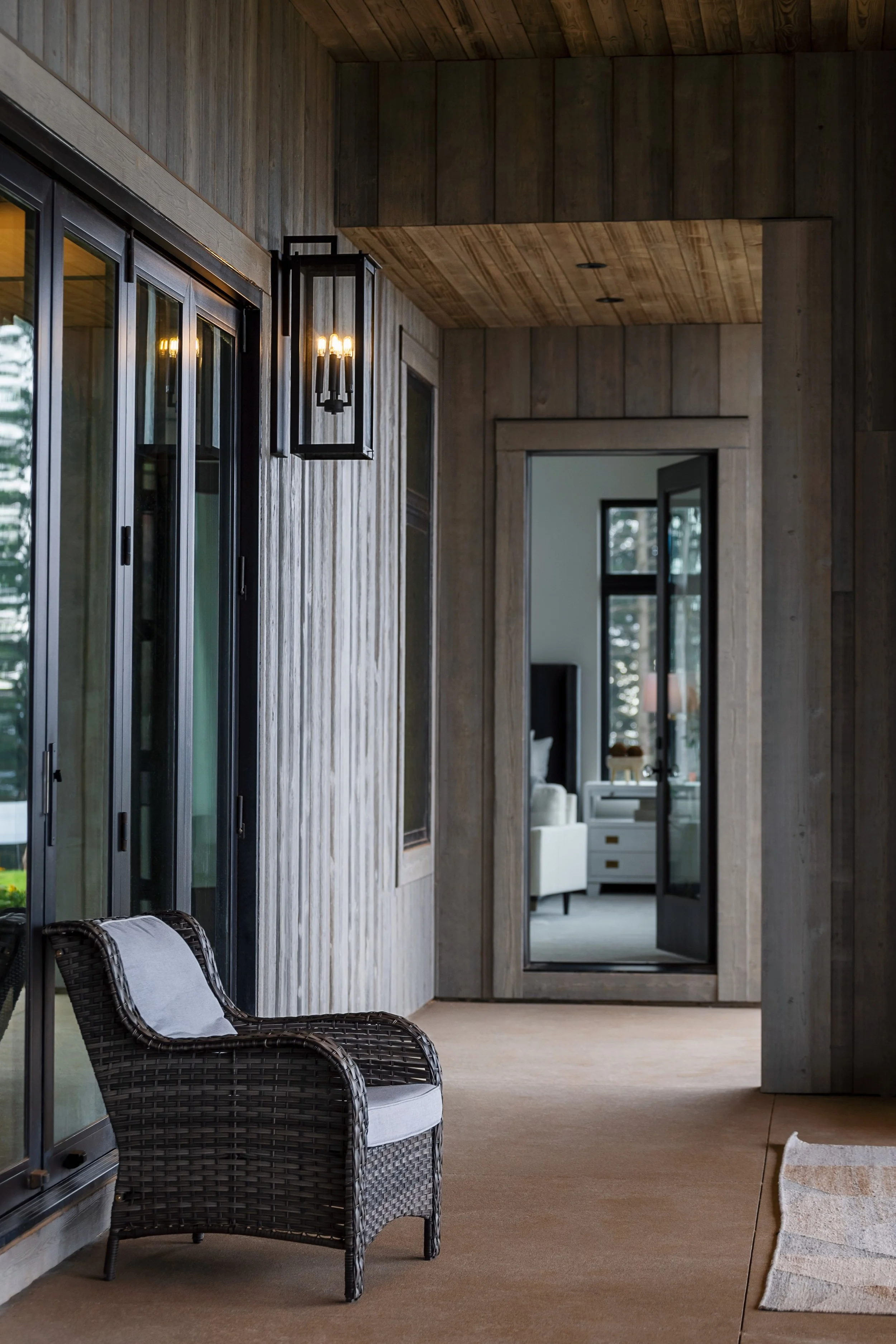 A cozy outdoor porch area with a wicker chair, sliding glass door, wooden walls, and a large mirror reflecting a room with a white chair and window.