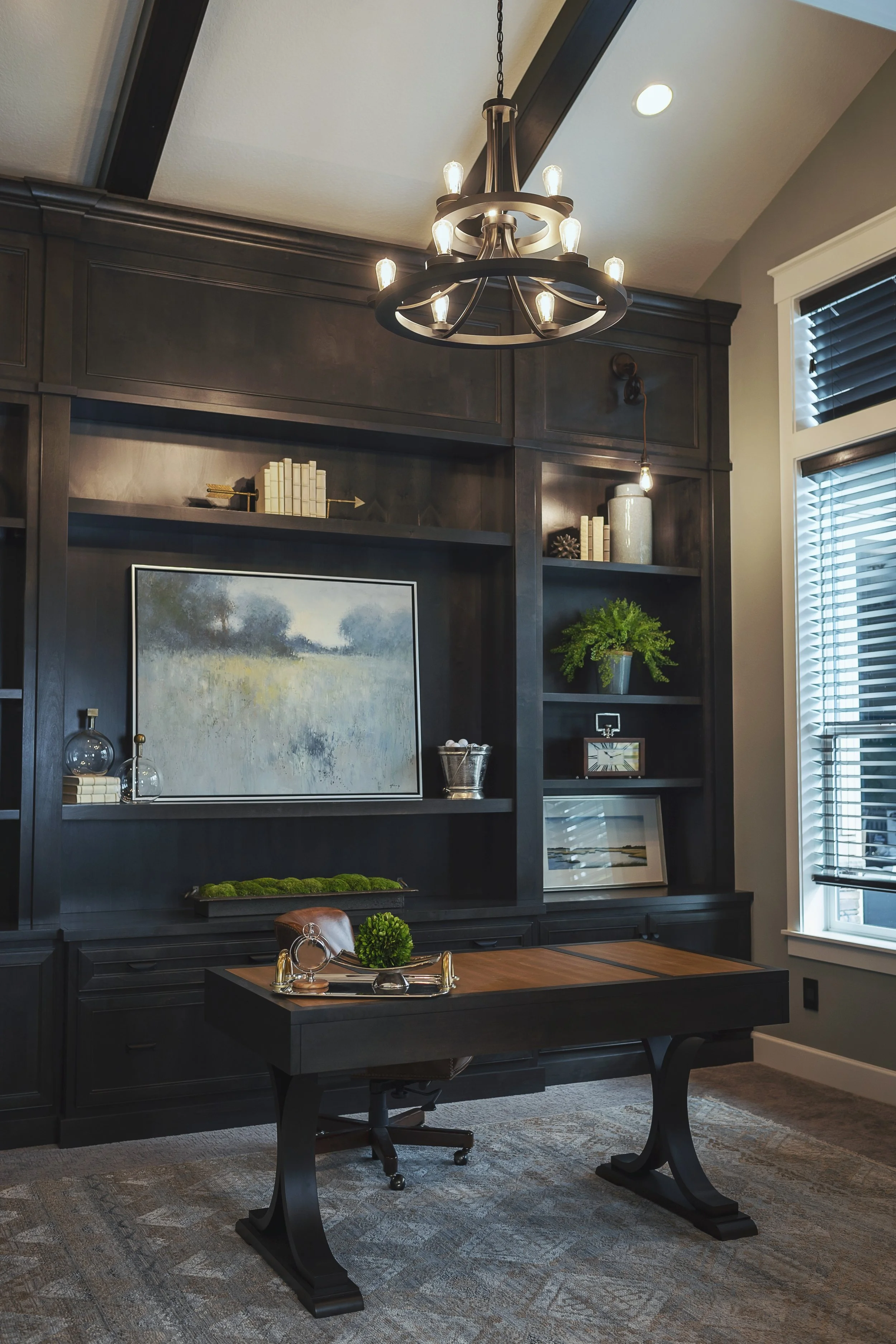 A well-lit home office with a black wooden desk, a black chair, a large dark wooden bookshelf, decorative items, and a chandelier with lights hanging from the ceiling near a window with blinds.