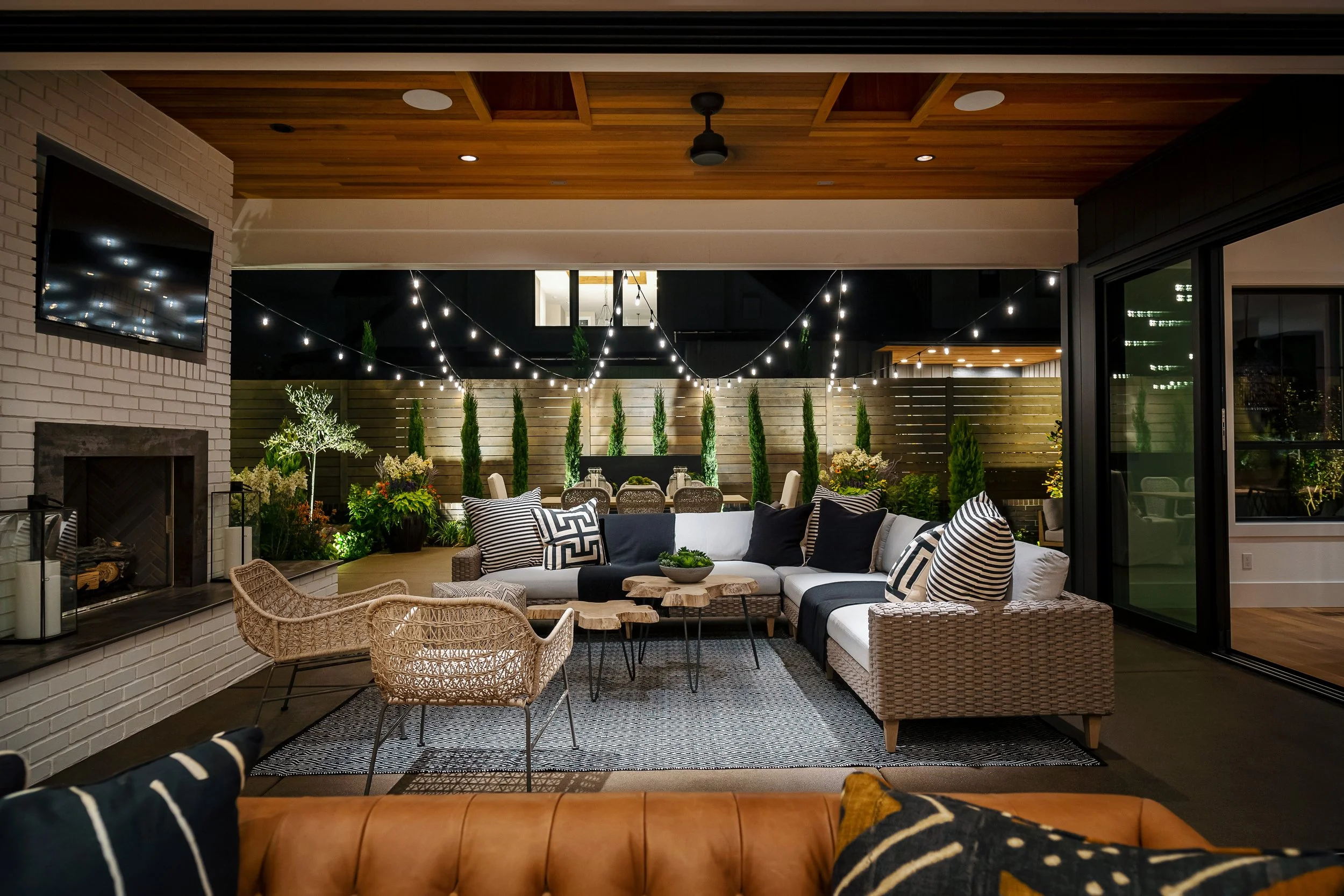 Outdoor patio with string lights, a white sectional sofa with black and white pillows, wicker chairs, a round wooden coffee table, plants, a brick fireplace, and a flat-screen TV mounted on the wall.