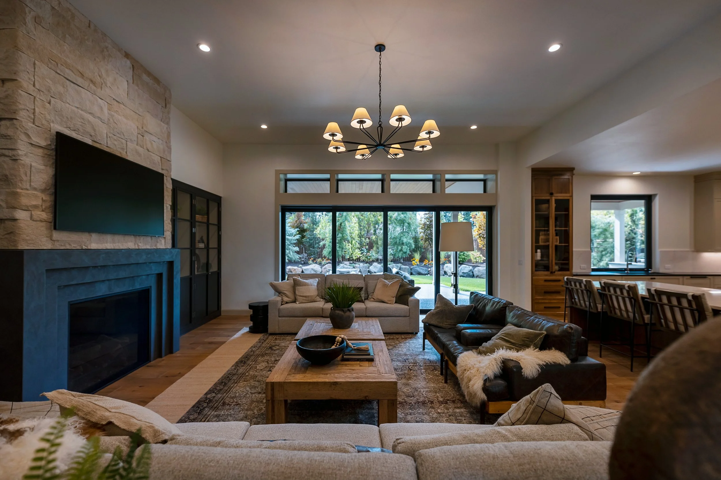 Bright living room with stone fireplace, large windows, wooden dining table, sofas, and modern chandelier with multiple lamps