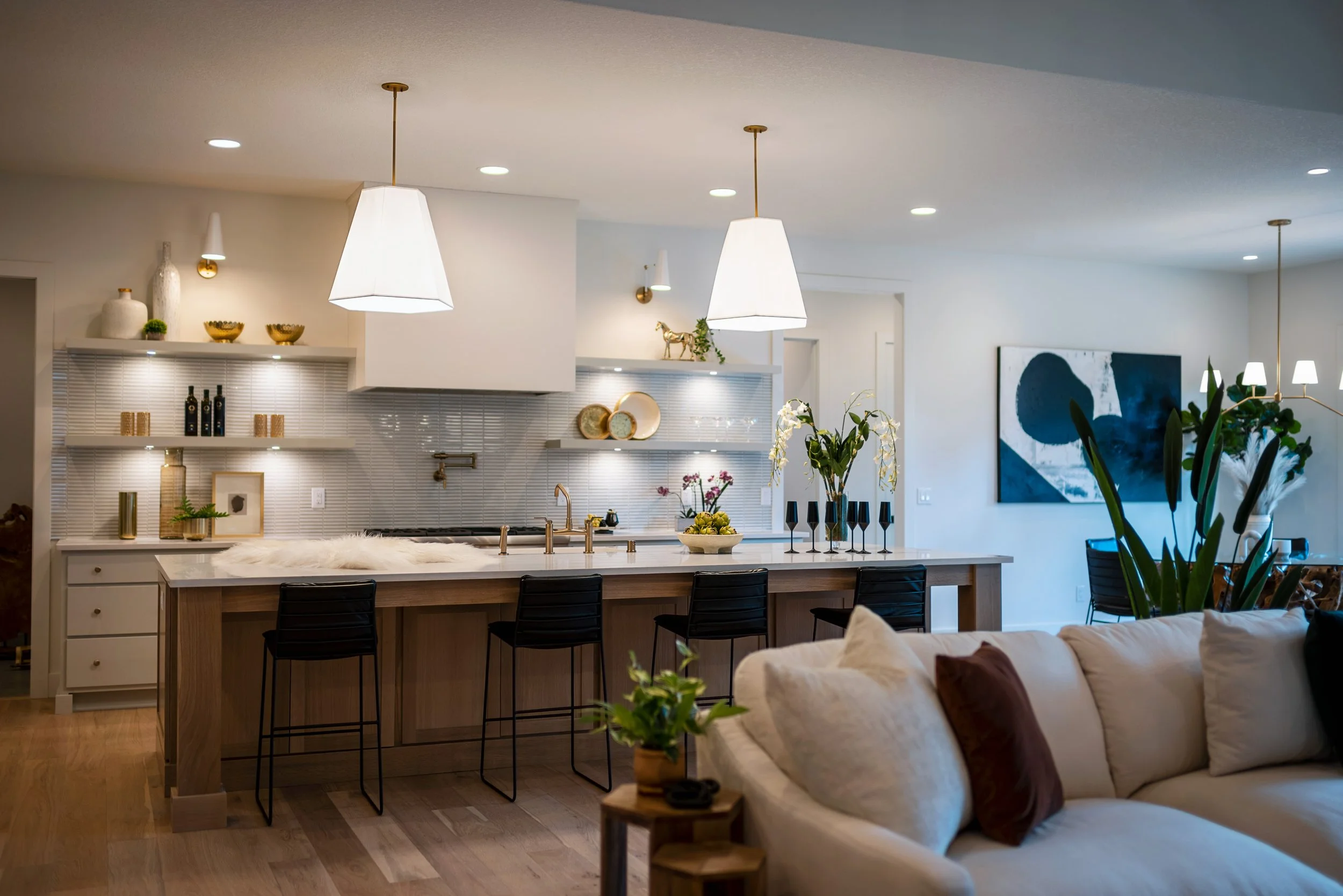 Modern kitchen and living room with white cabinets, gold accents, pendant and wall lights, black bar stools, green plants, and abstract artwork.
