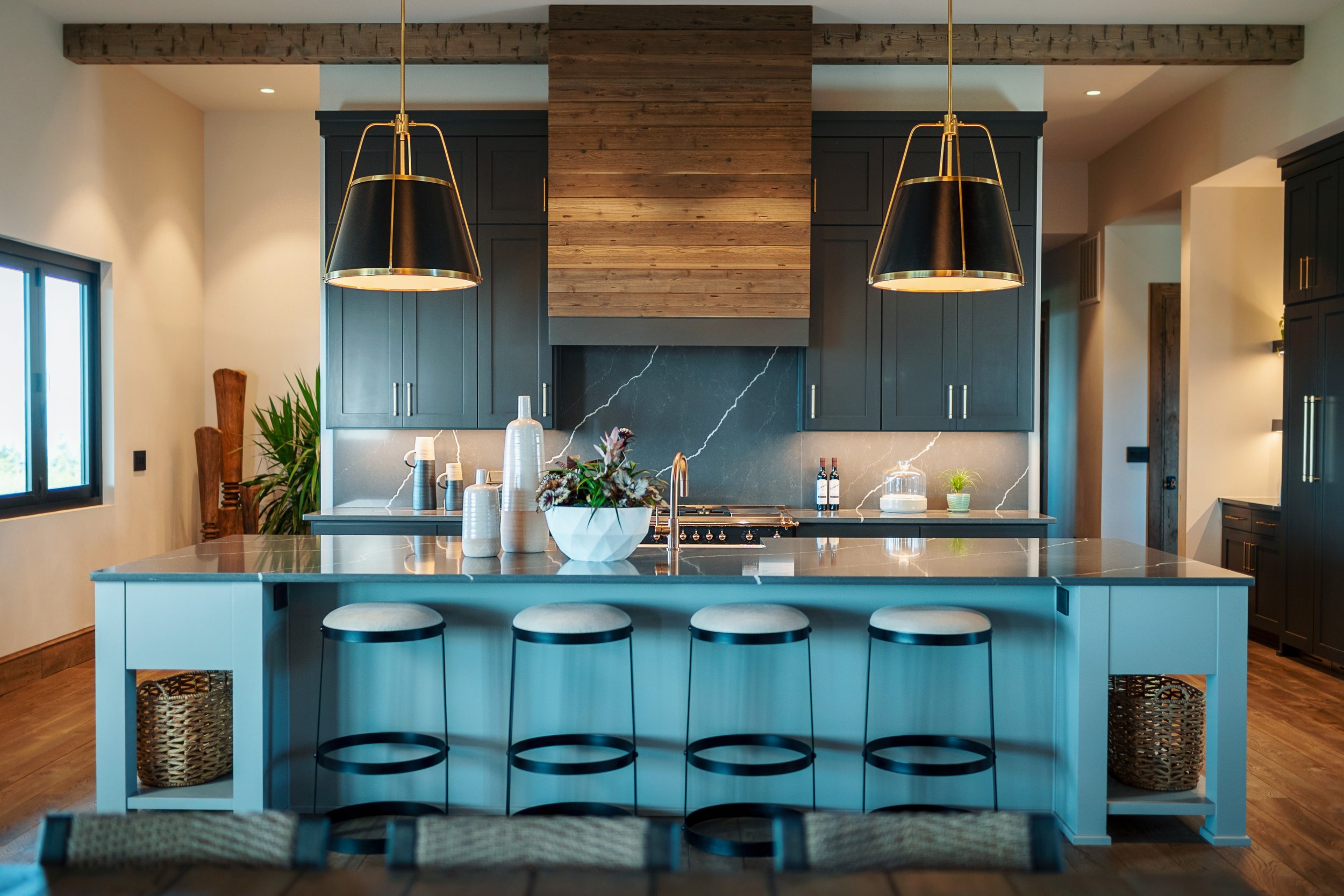 Modern kitchen with a large island in light blue, featuring five black barstools with round seats. The background includes black cabinetry, a slate backsplash with white veining, and a range hood. Two black and gold pendant lights hang above the isla