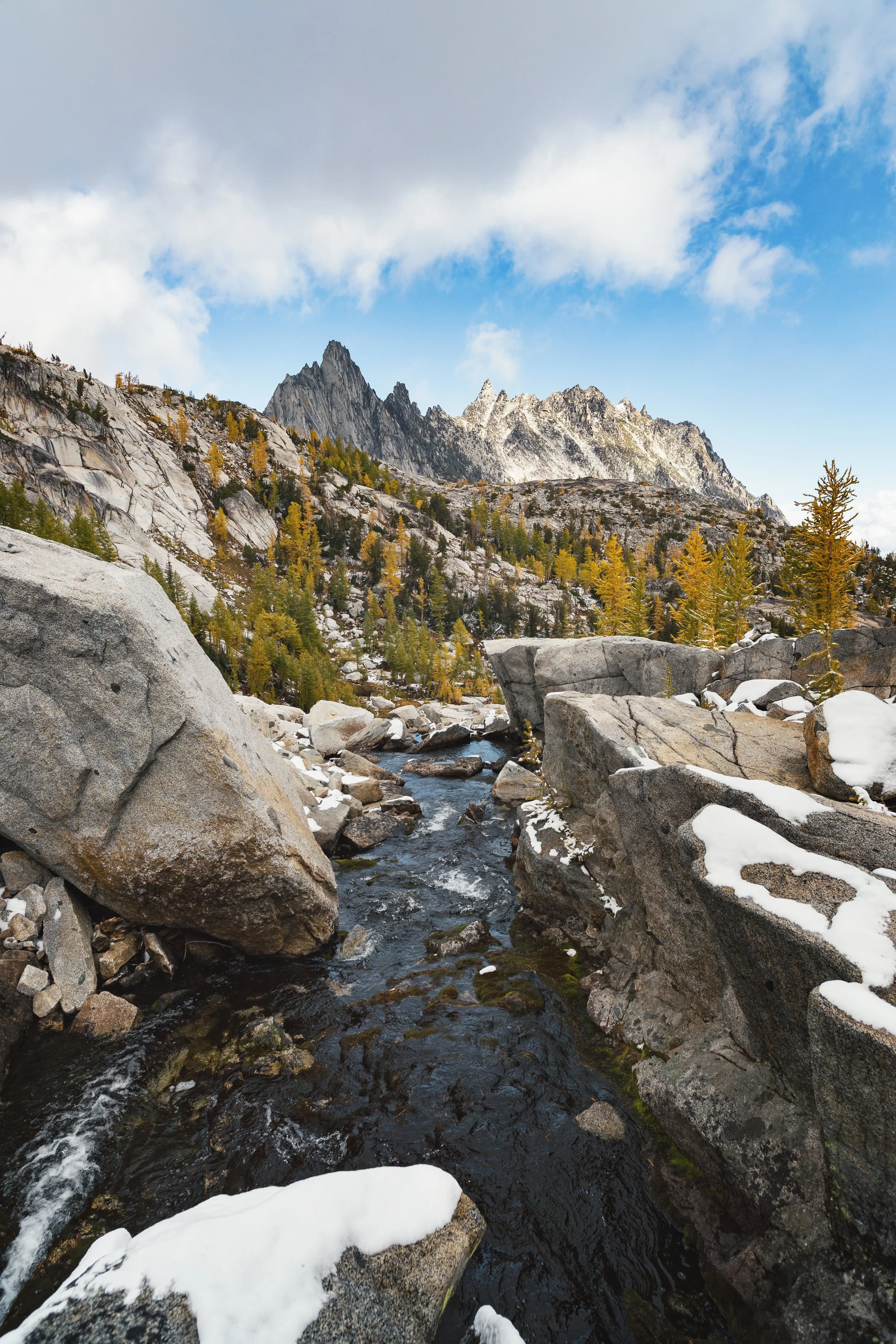 A mountain landscape with a flowing stream in the foreground, rocky terrain, trees with yellow leaves, and jagged snow-capped peaks in the background under a partly cloudy sky.