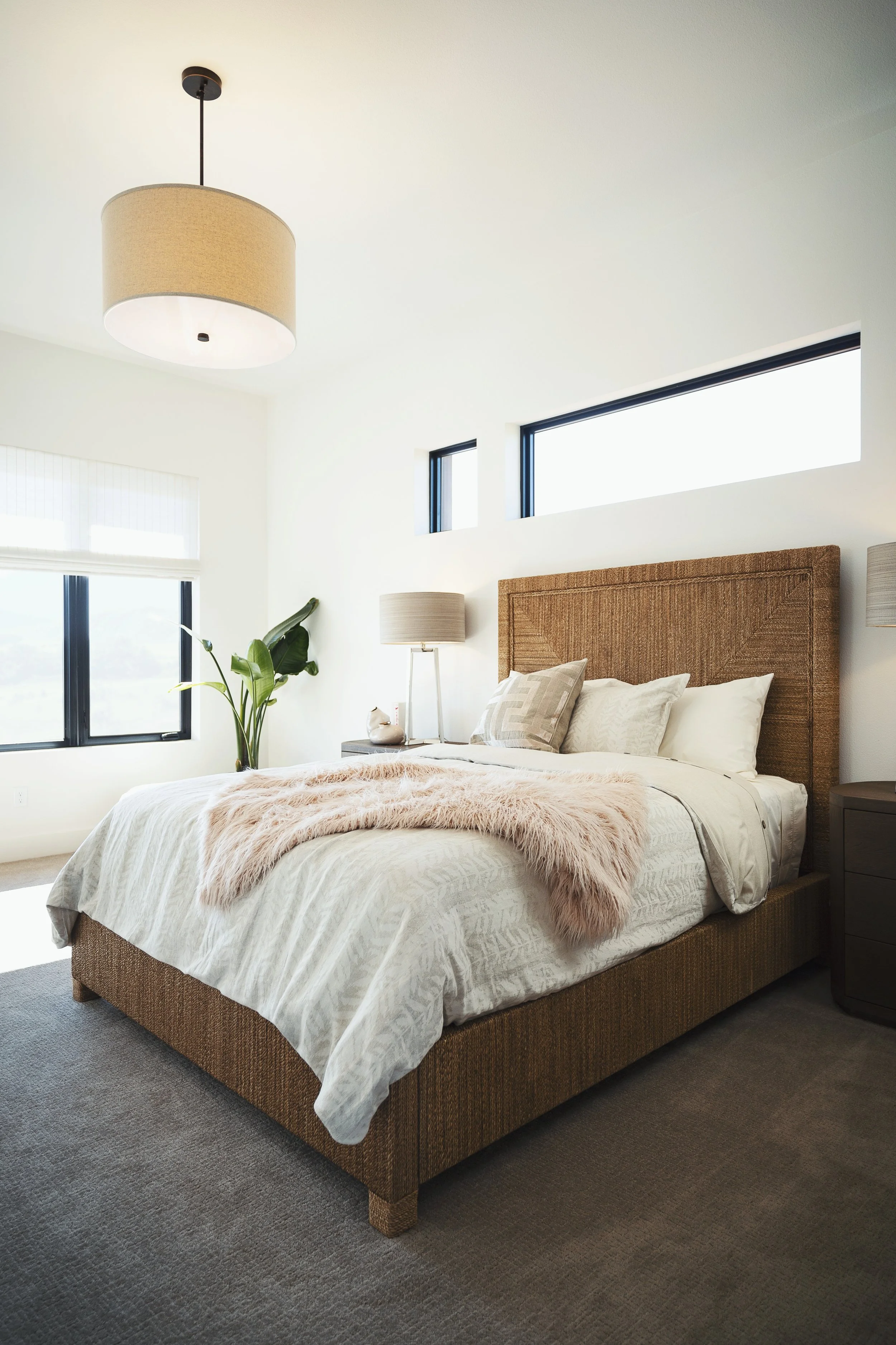 A neatly made bed with a beige woven headboard, white bedding, and decorative pillows, situated in a modern bedroom with large windows, a gray nightstand, a desk lamp, and a lush green plant.