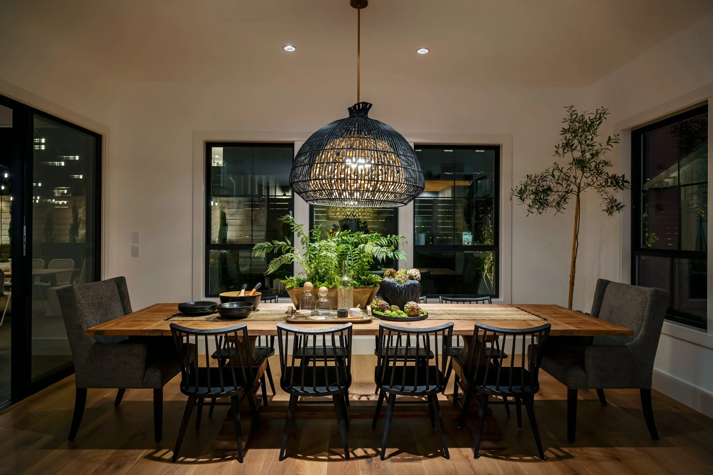 A dining room with a wooden table, black chairs, and a large black woven pendant light hanging above. The table has plants, bowls, and decorative items on it. There are large windows behind the table and an indoor plant to the right. The room has a m