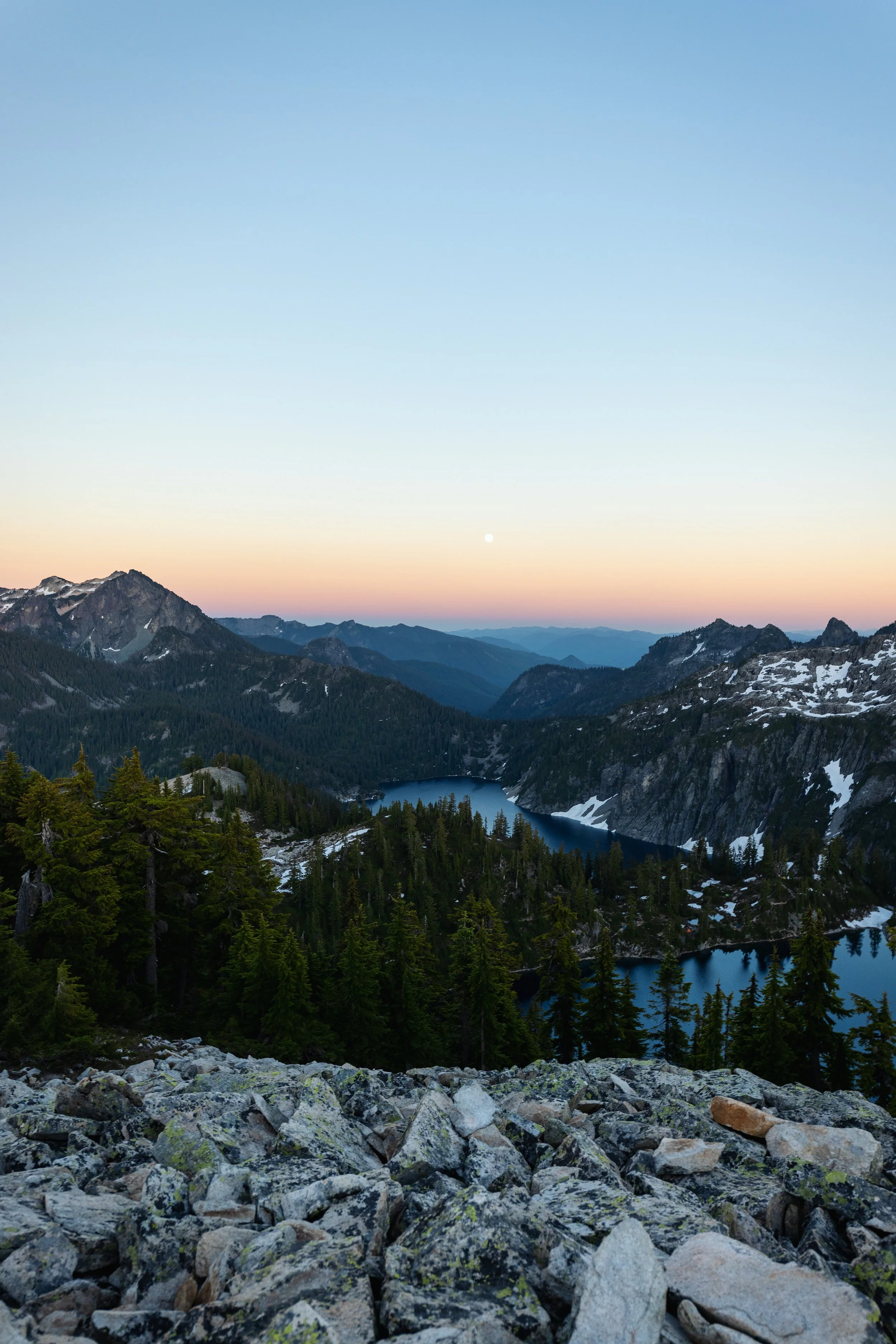 Mountain landscape at sunrise with rocky foreground, dense pine trees, mountain peaks with some snow, a lake, and a clear sky with the moon visible.
