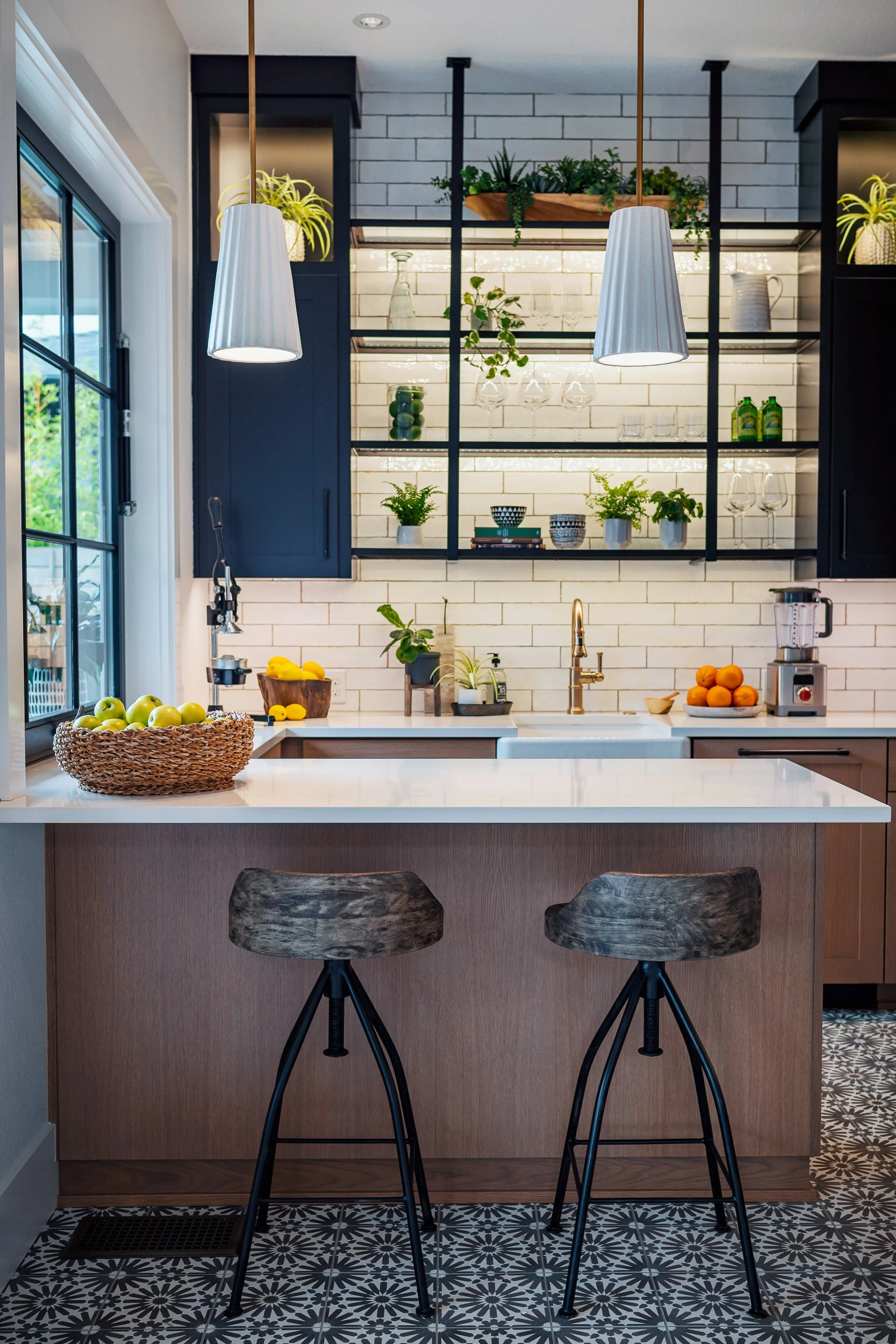 Modern kitchen with black pendant lights, open shelving with plants and glassware, white subway tile backsplash, and a kitchen island with two wooden bar stools, patterned tile floor, and fruit basket.