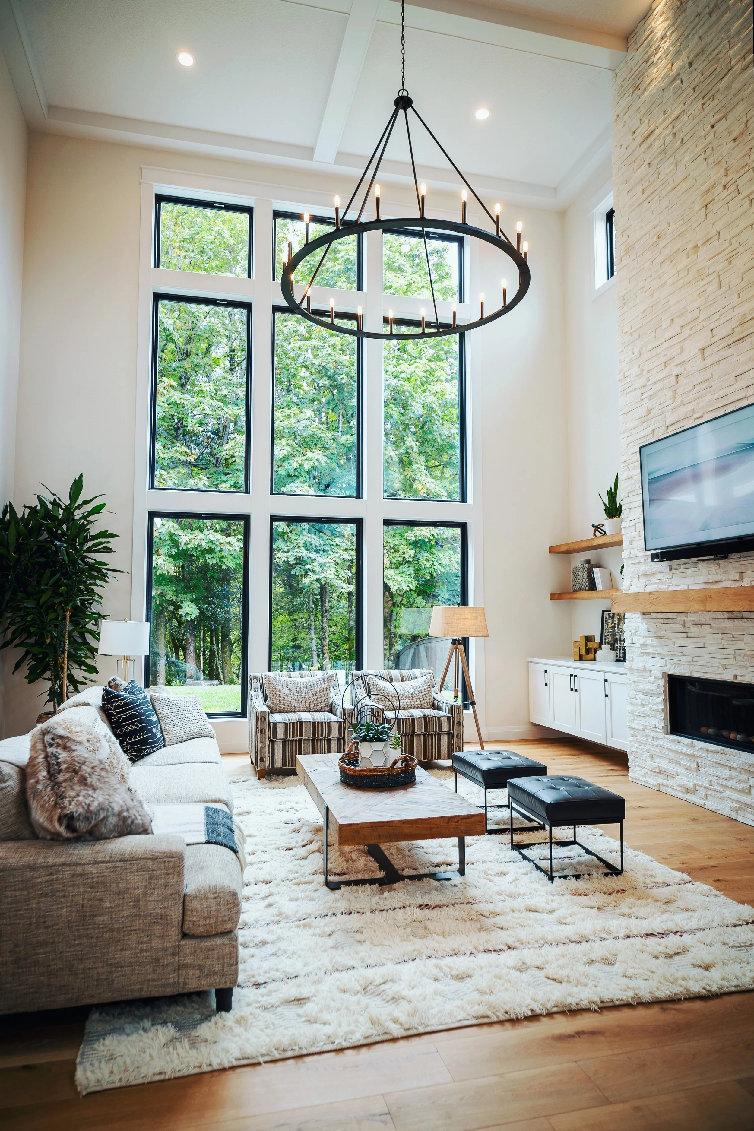 Living room with large window showing green trees outside, modern chandelier, cream-colored sofa with patterned pillows, striped armchair, wooden coffee table with plants, TV mounted on a stone wall, white cabinets, and various decor items.