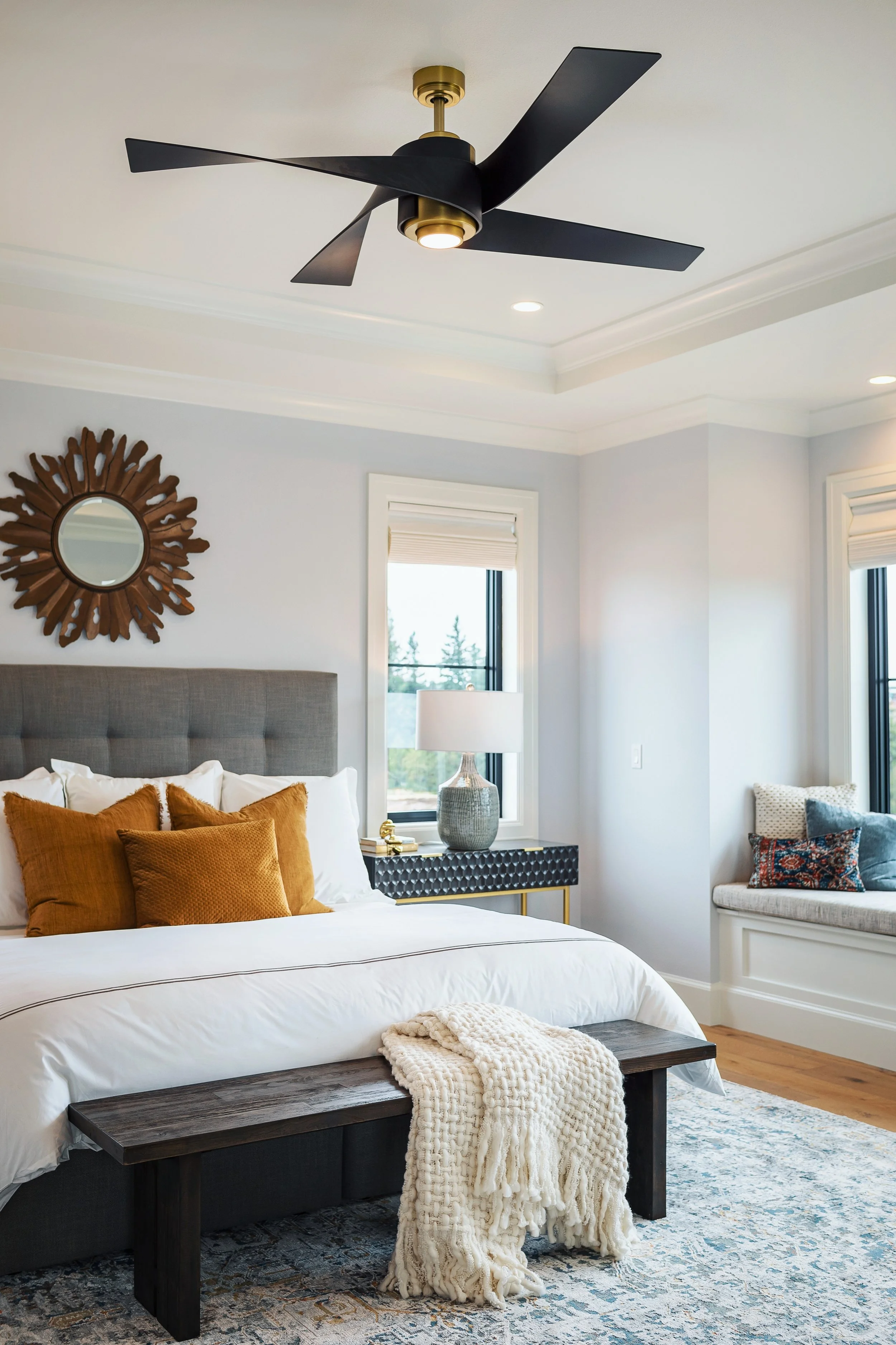 Bedroom with a modern ceiling fan, a bed with brown and white pillows, a dark wood bench at the foot of the bed with a cream blanket, a blue patterned area rug, a window with white trim, and a side table with a lamp and decorative vase.