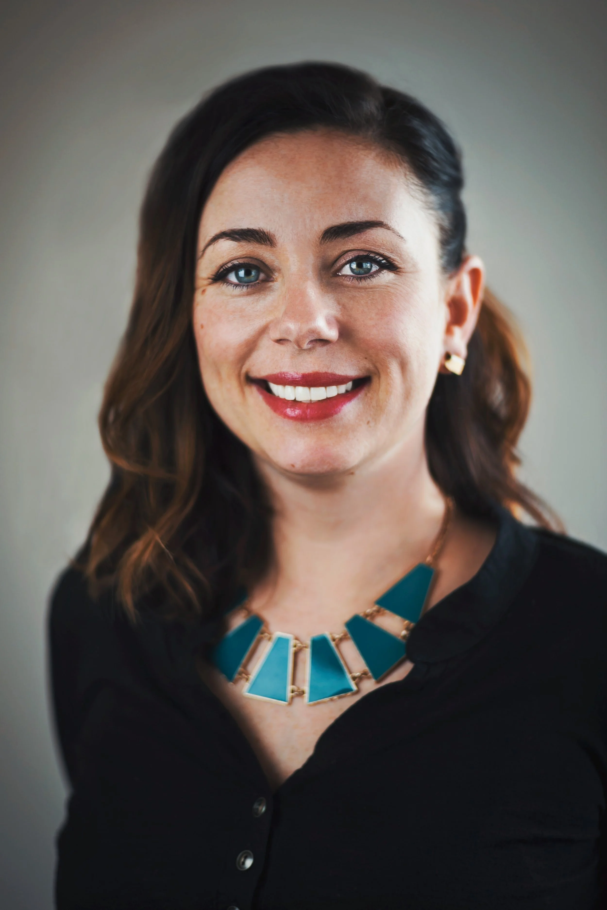 A woman with dark hair and blue eyes, smiling, wearing a black top, a turquoise geometric necklace, and gold earrings.