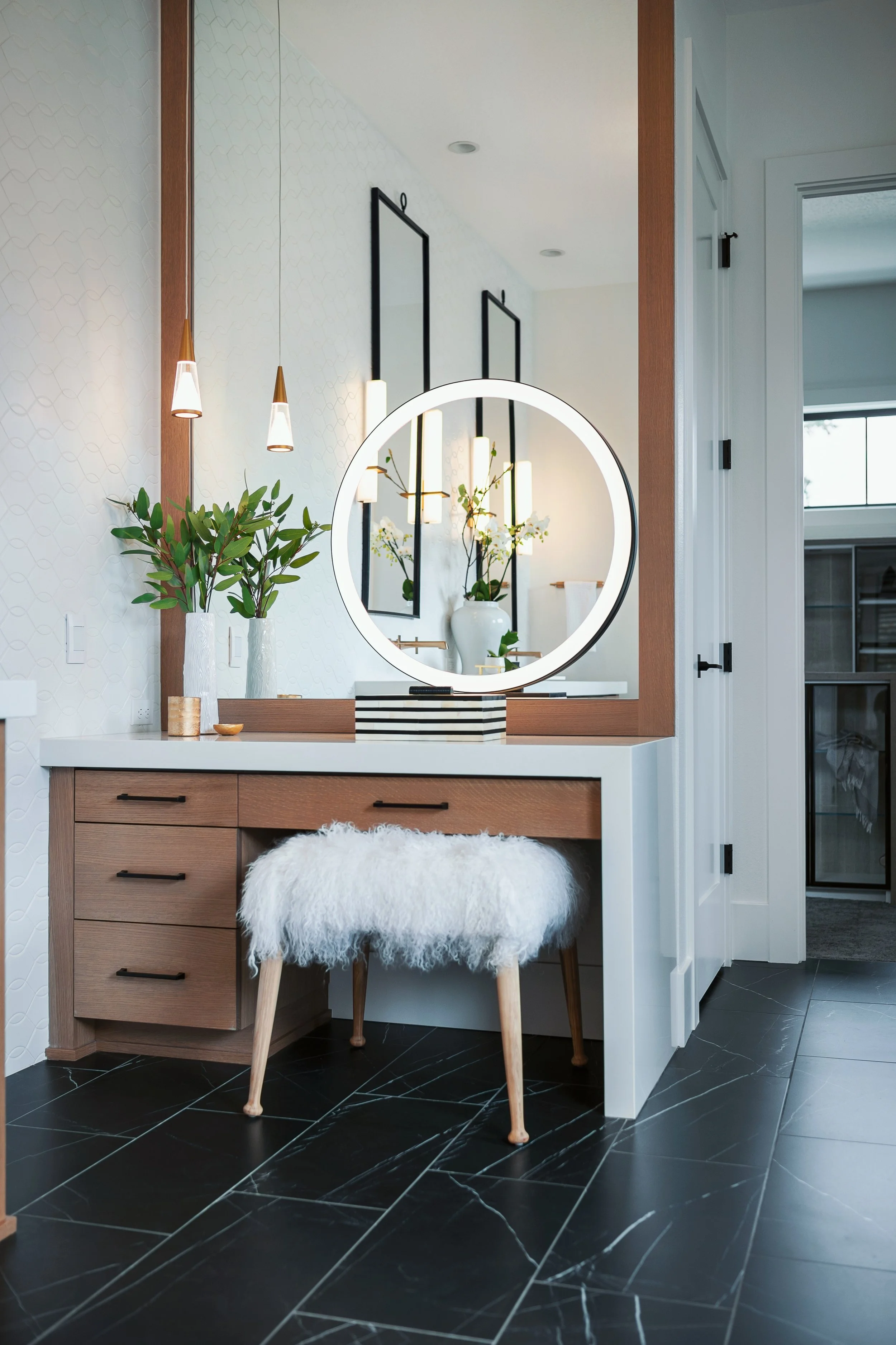 A modern vanity area with a round illuminated mirror, two vases with green leaves, and a fluffy white stool, in a room with dark tile flooring and white walls.