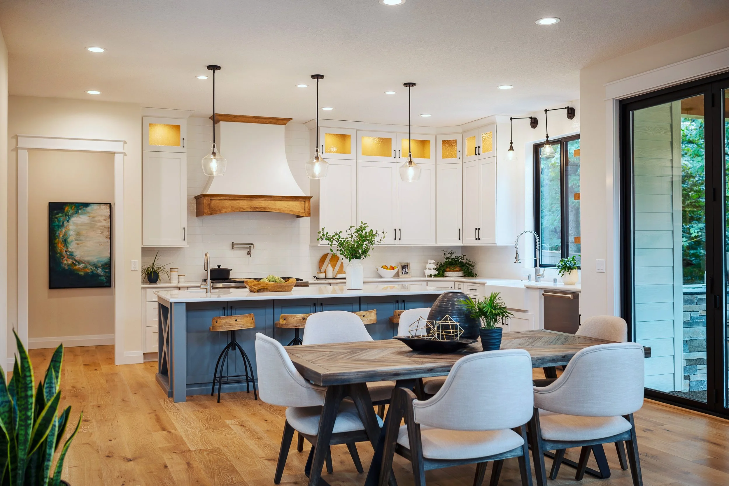 A modern kitchen with white cabinets, a farmhouse sink, and a blue kitchen island, overlooking a dining area with a wooden table and white upholstered chairs, illuminated by pendant lights and natural light from large sliding glass doors.