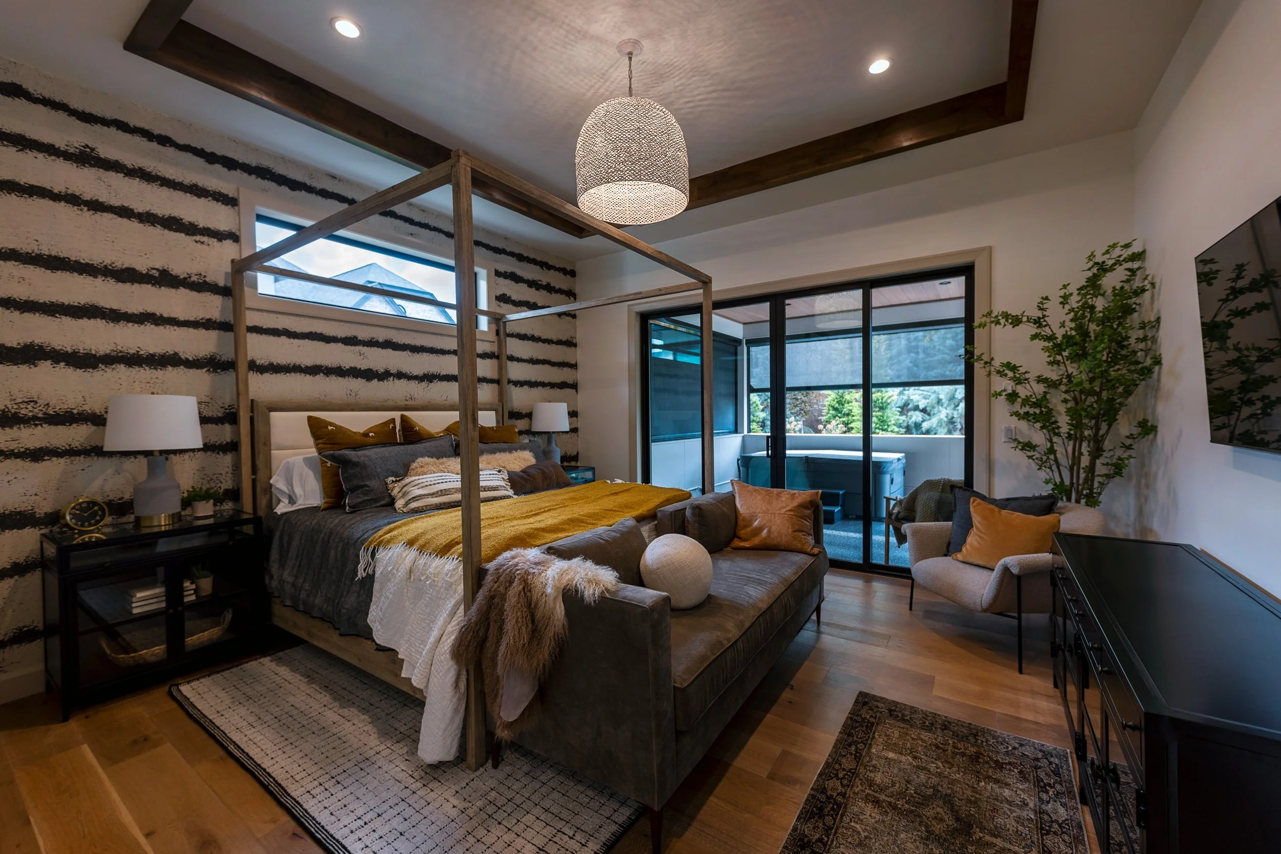 Interior of a modern bedroom with a wooden four-poster bed, beige and brown tones, textured black and white striped wall, sliding glass doors leading to a balcony, and a seating area with armchairs, pillows, and indoor plants.