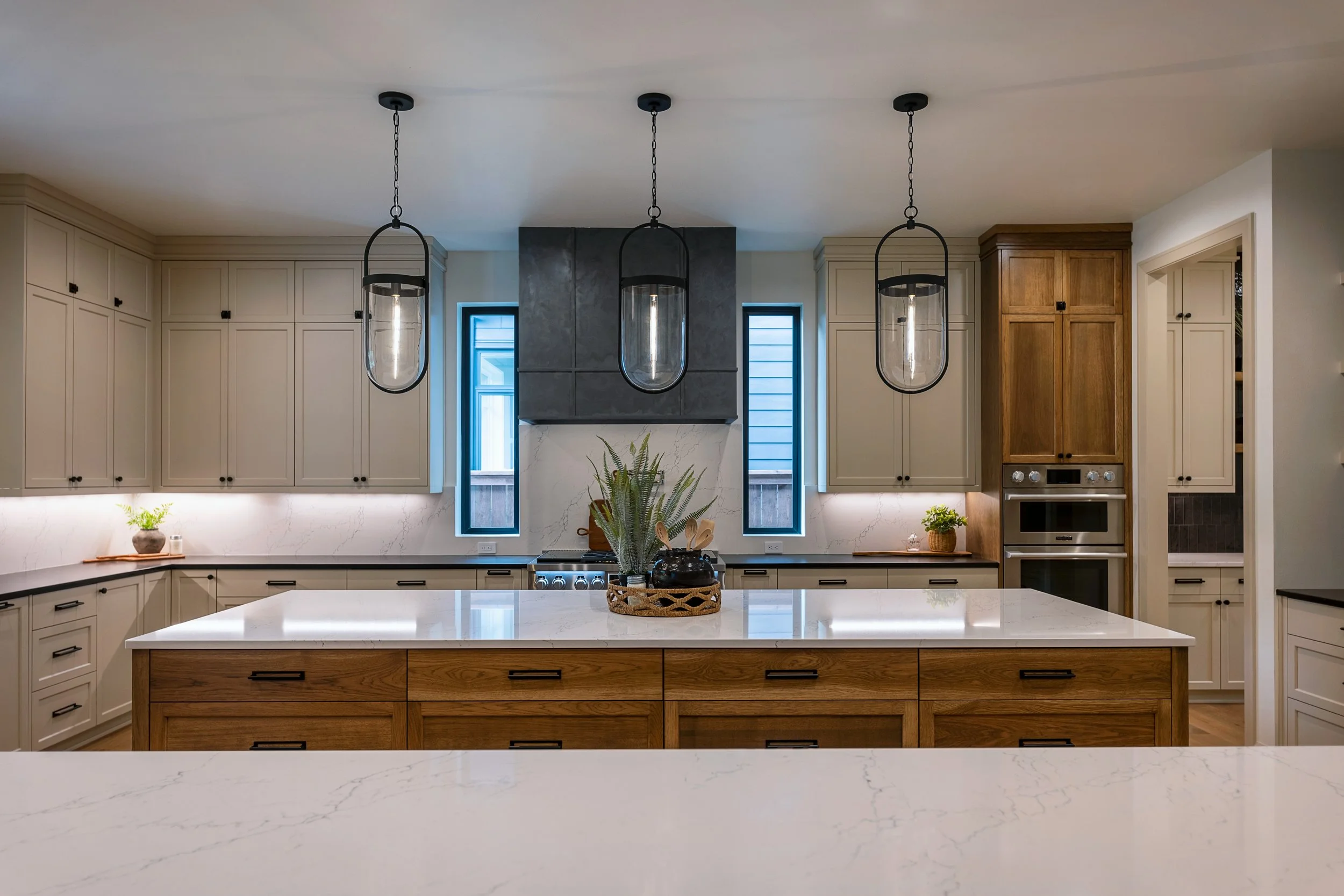 Modern kitchen with white cabinetry, black hardware, a large white marble island, a stainless steel oven, wooden accents, pendant lights, and small plants on the countertops.