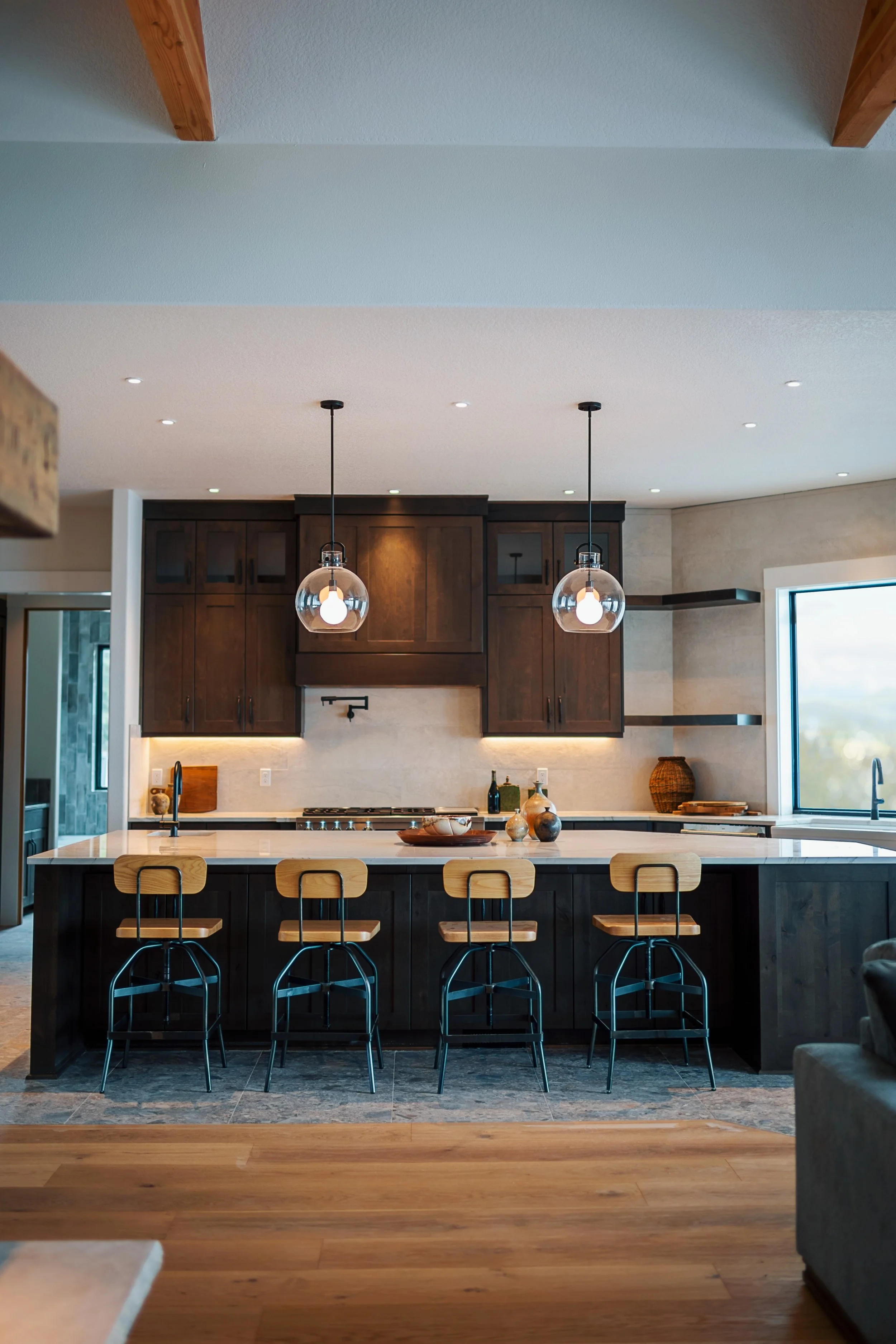 Modern kitchen with dark wood cabinets, white countertop, pendant lights, and bar stools at island, large window, and wooden flooring.