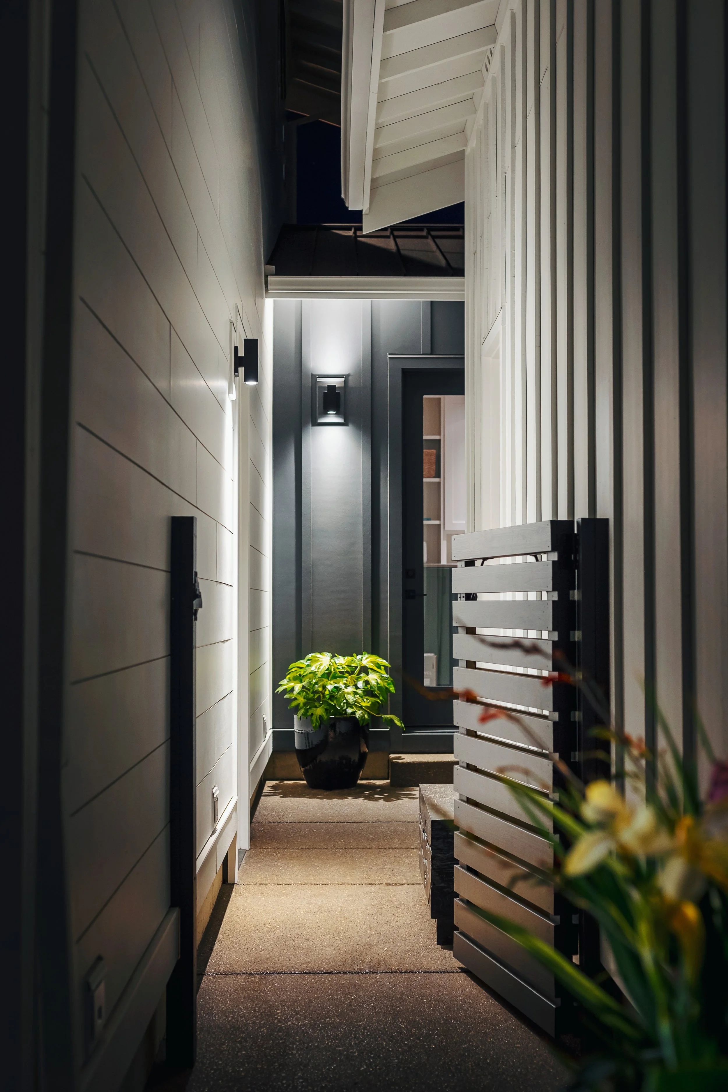 A narrow outdoor corridor at night with a potted plant at the end, wooden slat fence on the right, white siding walls on the left, and dark door at the end of the corridor.