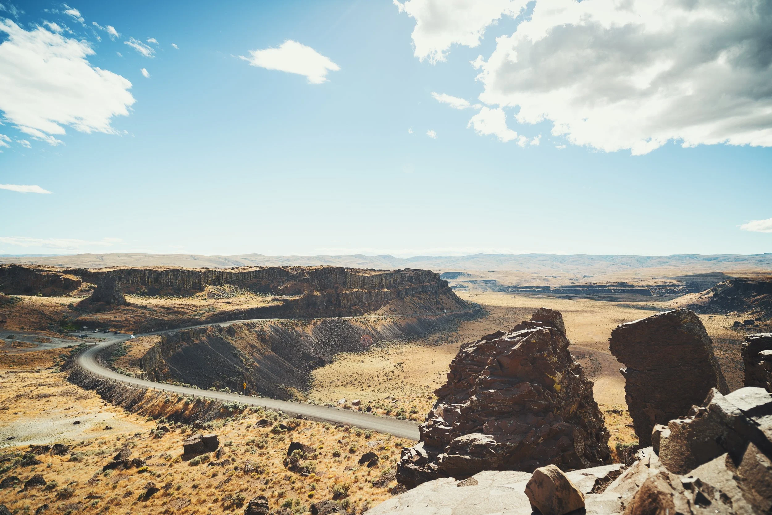Scenic view of a winding road through a desert landscape with rocky formations and distant hills, under a partly cloudy sky.