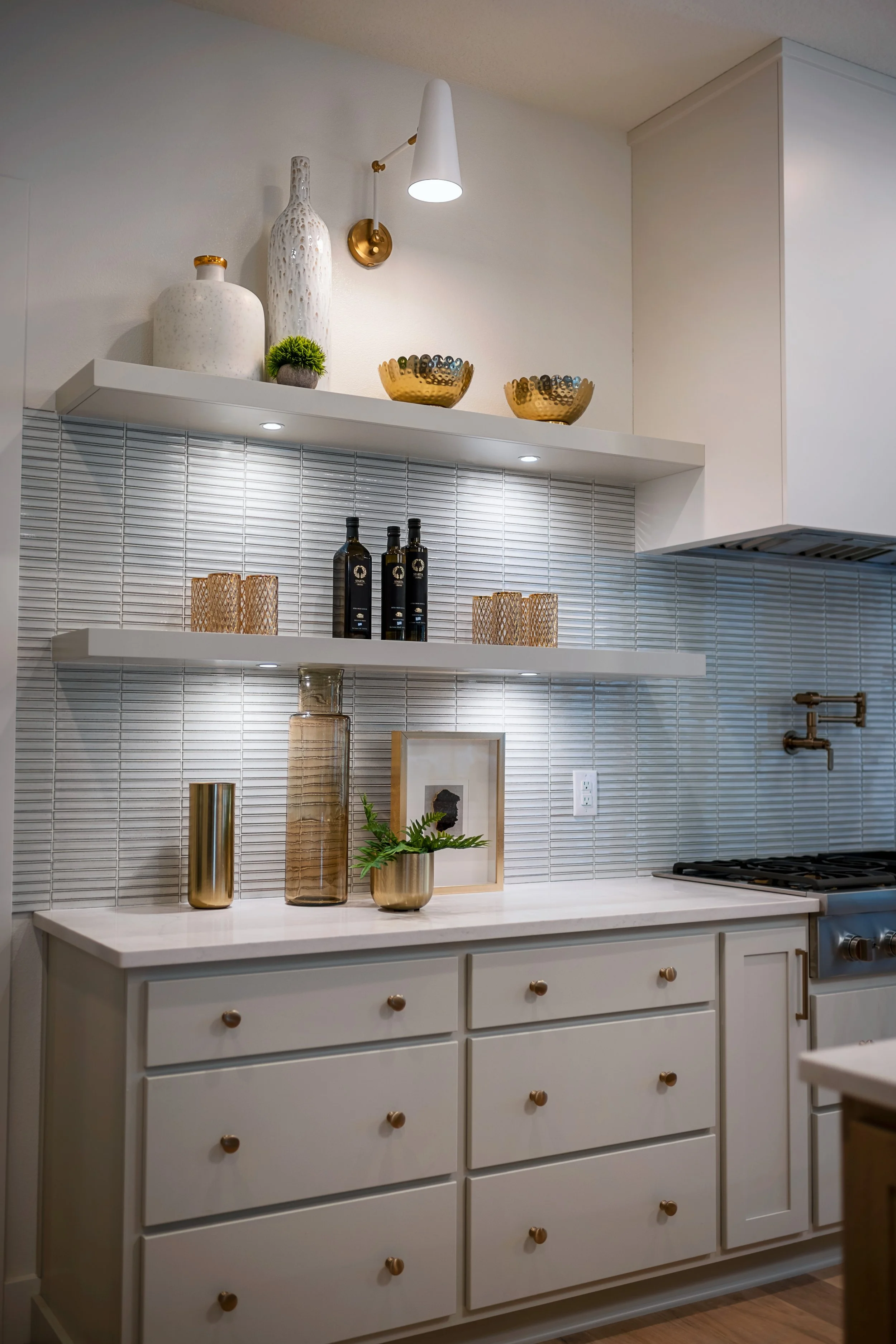 A modern kitchen with white cabinetry, gold knobs, three drawers, decorative vases, bottles, bowls, and plants on white shelves with tap and stove in the background.