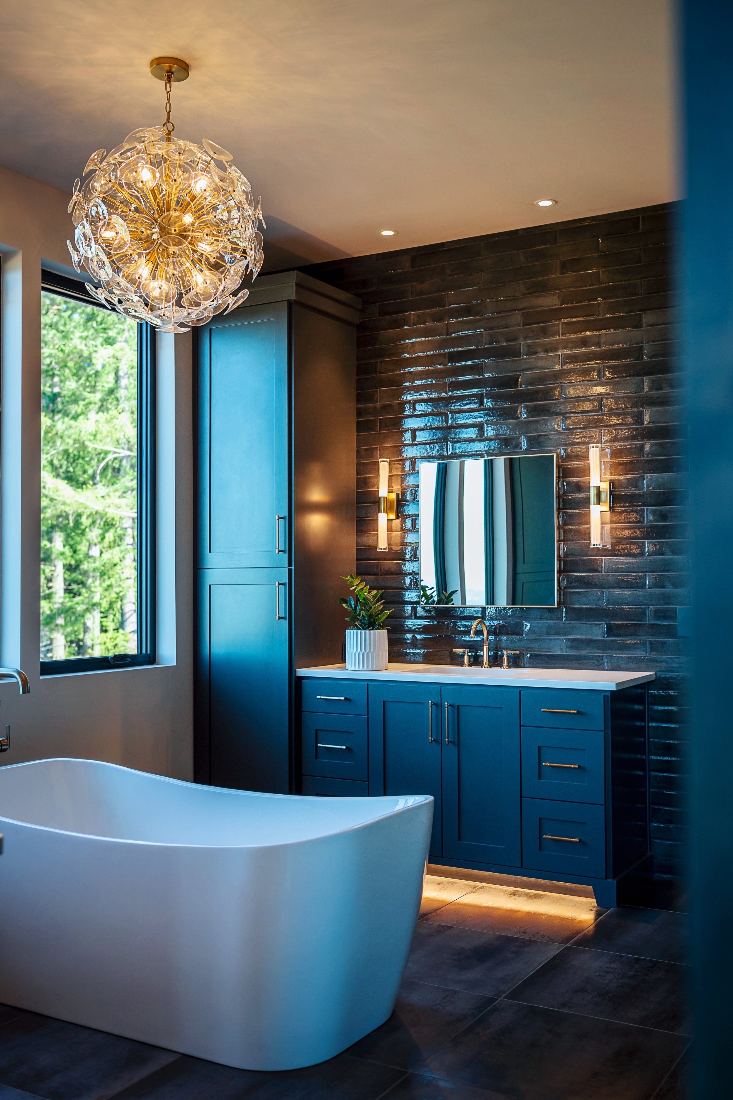 A modern bathroom with a white bathtub, blue vanity cabinets, a round mirror, black brick wall, window with view of greenery, decorative plant, chandelier with glass elements, and warm lighting.