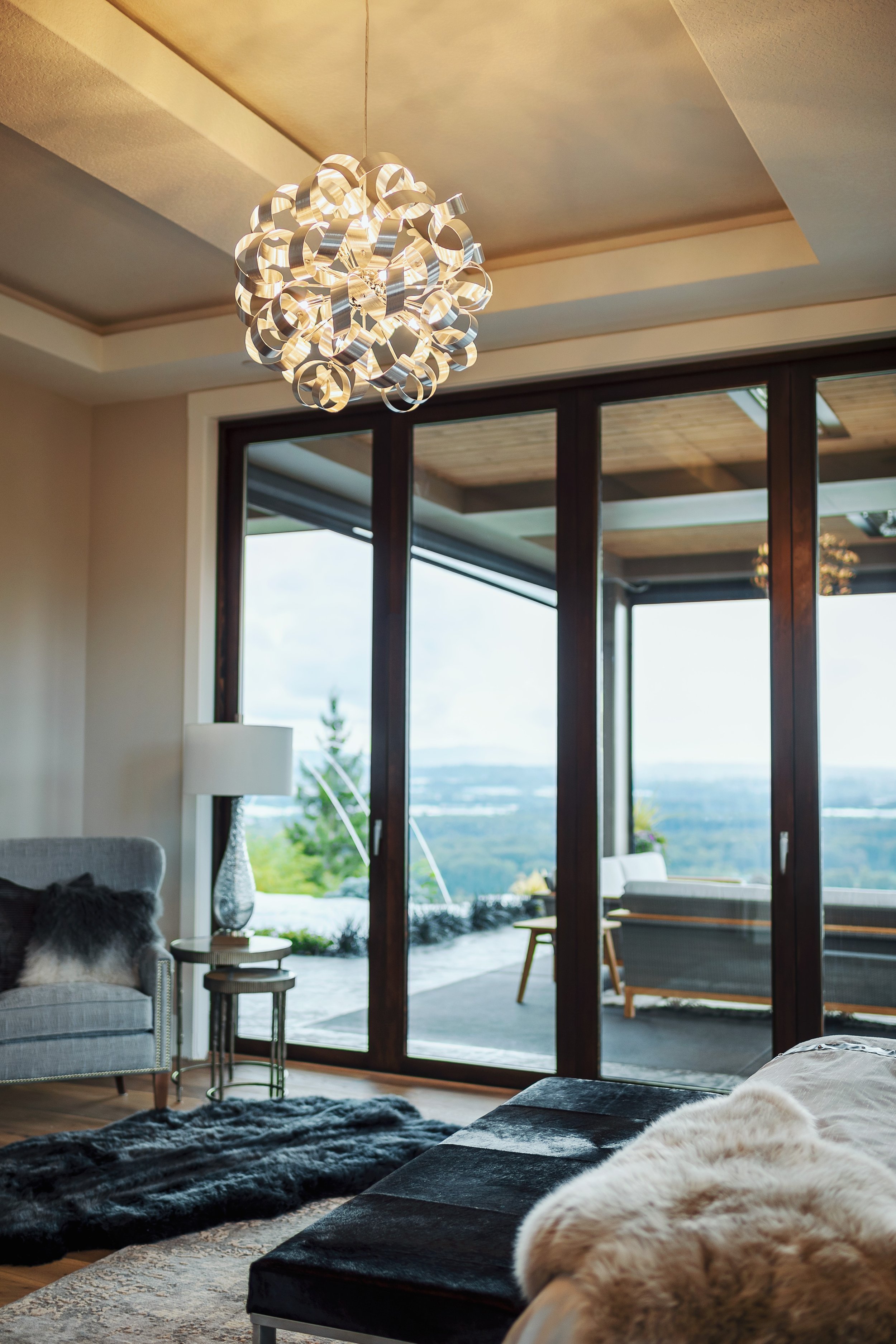 Living room with modern chandelier, sliding glass doors opening to an outdoor patio, with outdoor furniture and view of trees and water in the distance.